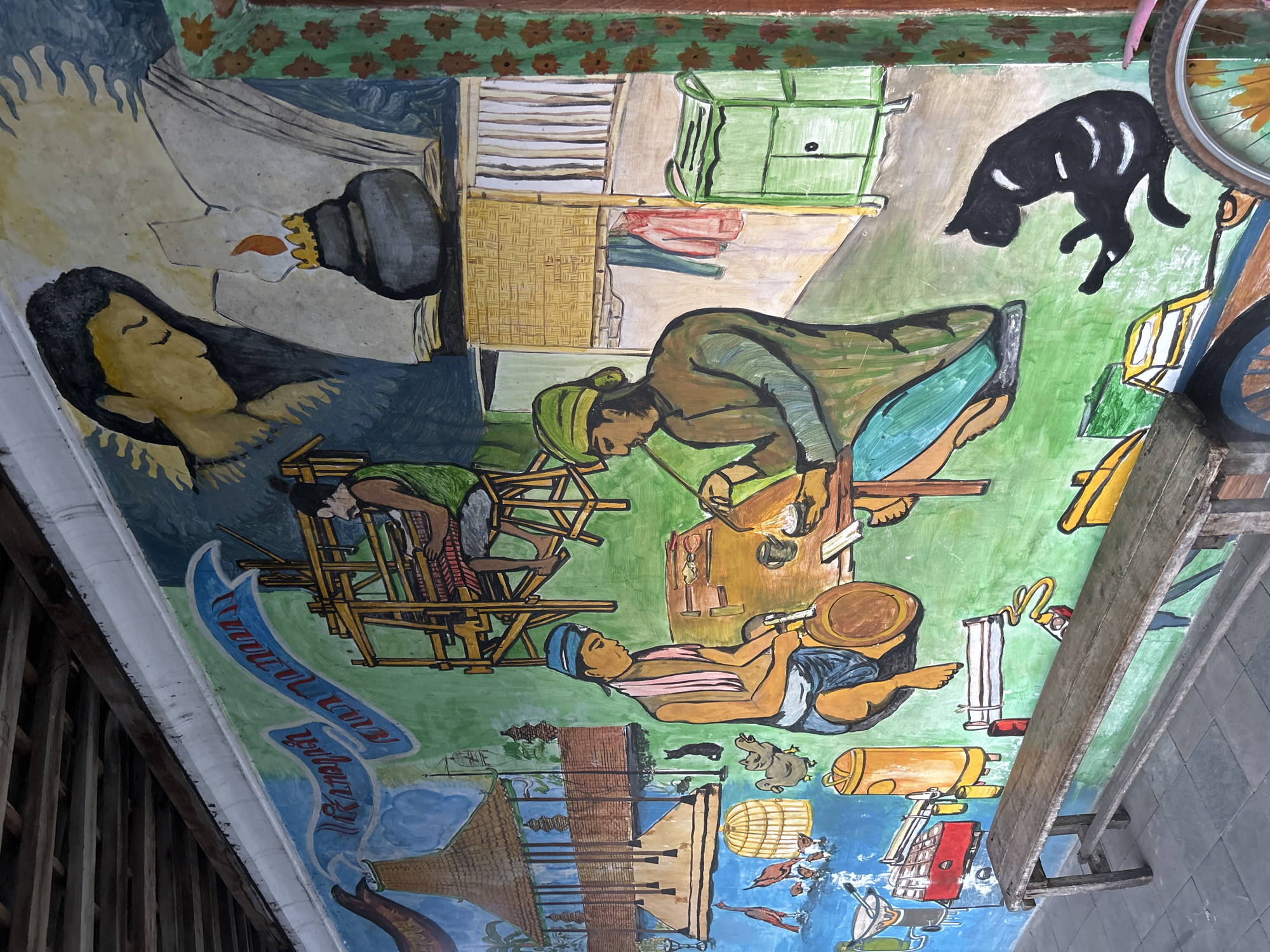 This is a colorful, somewhat naive mural painted on what appears to be a ceiling or upper wall, likely in Yogyakarta, Indonesia. The mural depicts scenes of daily life and rural activities.
In the upper left, a sleeping figure with dark hair and yellow skin is partially visible, with a hand reaching towards them holding a black object with a golden crown. Above this, a border of brown floral designs on a green background runs across the top.
Below the sleeping figure, a person in green is bent over a large, wooden loom-like structure, appearing to be engaged in weaving or textile work. To their right, another person in brown and green is hunched over a workbench, possibly crafting or repairing something, with tools visible. Further to the right, a person in a blue headscarf and pink sash is lying down, holding a tool or object, with a large barrel beside them.
The background is a vibrant green, suggesting an outdoor or natural setting. Scattered throughout the scene are various objects, including woven baskets, green cabinets, red and blue fabrics, yellow containers, a birdcage, and several birds. A black cat is depicted in mid-leap in the upper right section. Part of a bicycle wheel is visible at the far top right.
In the lower portion of the mural, there are depictions of houses with thatched roofs, a body of water, and more stylized elements that could represent boats or structures. A blue ribbon with white lettering runs diagonally through the lower left of the image, containing text that appears to be in Javanese script. The overall impression is one of a lively and industrious community. The mural's style suggests a folk art or traditional artistic approach. The orientation of the image suggests it is looking upwards.