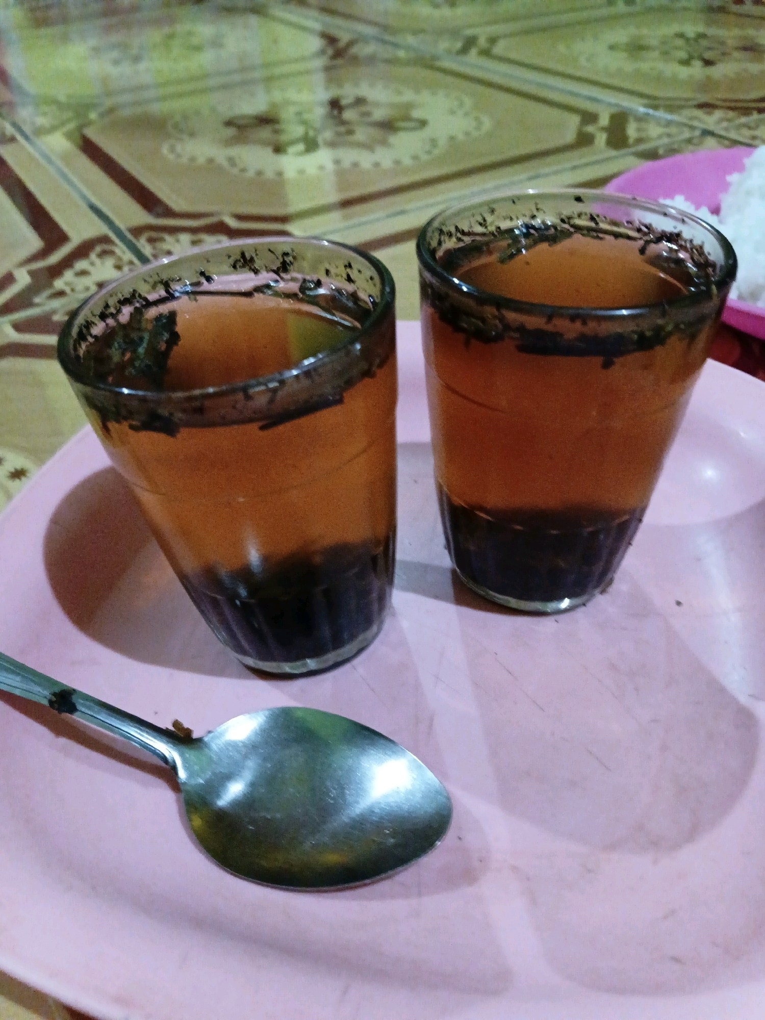 Two glasses of brown liquid with dark sediment at the bottom and on the rims are placed on a pink plate. A metal spoon rests on the plate in front of the glasses. In the background, a tiled surface with a patterned design is visible. A pink tray with white rice is partially visible on the right side of the image. The setting appears to be indoors, possibly in a residential area in Margo Rukun, Indonesia.