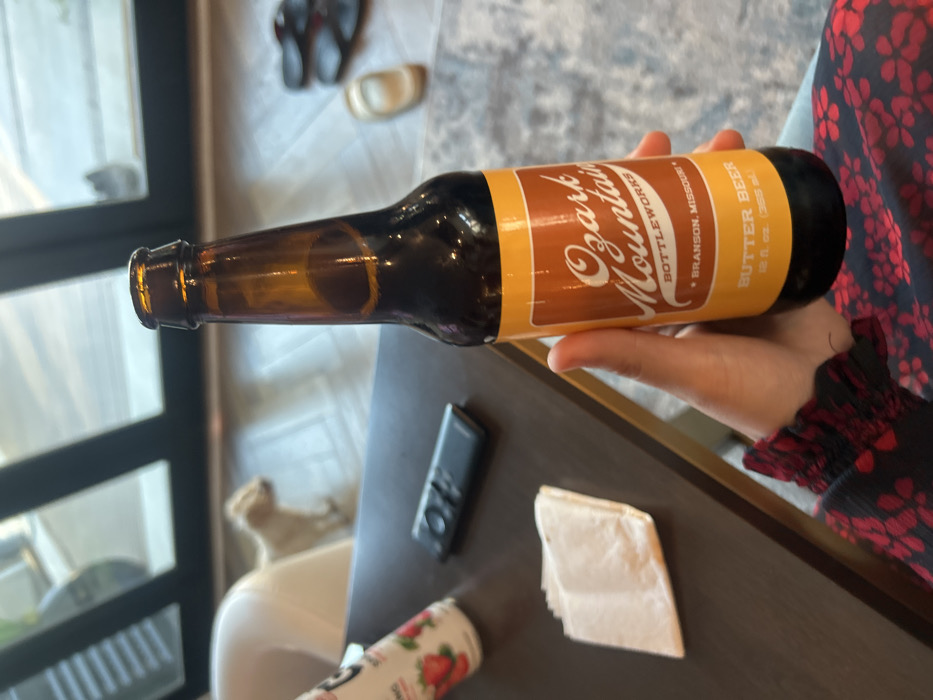 A nearly empty amber glass bottle labeled "Ozark Mountain Bottleworks Butter Beer, Branson, Missouri" rests horizontally in a hand. The label features a brown and cream color scheme. Below the bottle is a table with a remote control, a folded napkin, and a drink container decorated with strawberries. In the background is a window and a glimpse of slippers. The person holding the bottle wears a red and black floral print top.