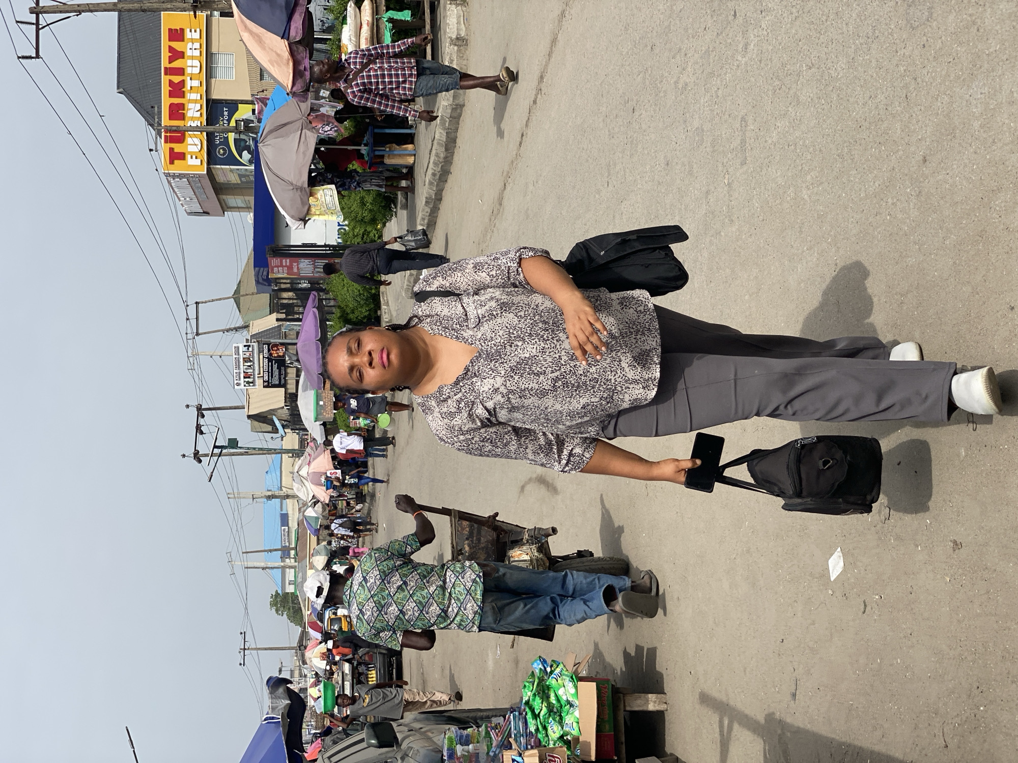 The image depicts an urban street scene in Lekki, Nigeria. A woman stands prominently in the foreground, holding a smartphone and a black shoulder bag. The street is crowded with pedestrians, vendors, and various objects associated with a bustling marketplace. Umbrellas and canopies provide shade for several stalls. A man with a cart is seen pulling it. A sign for "Türkiye Furniture" is visible in the background. The environment suggests a vibrant commercial area with a mixture of businesses and street vendors.