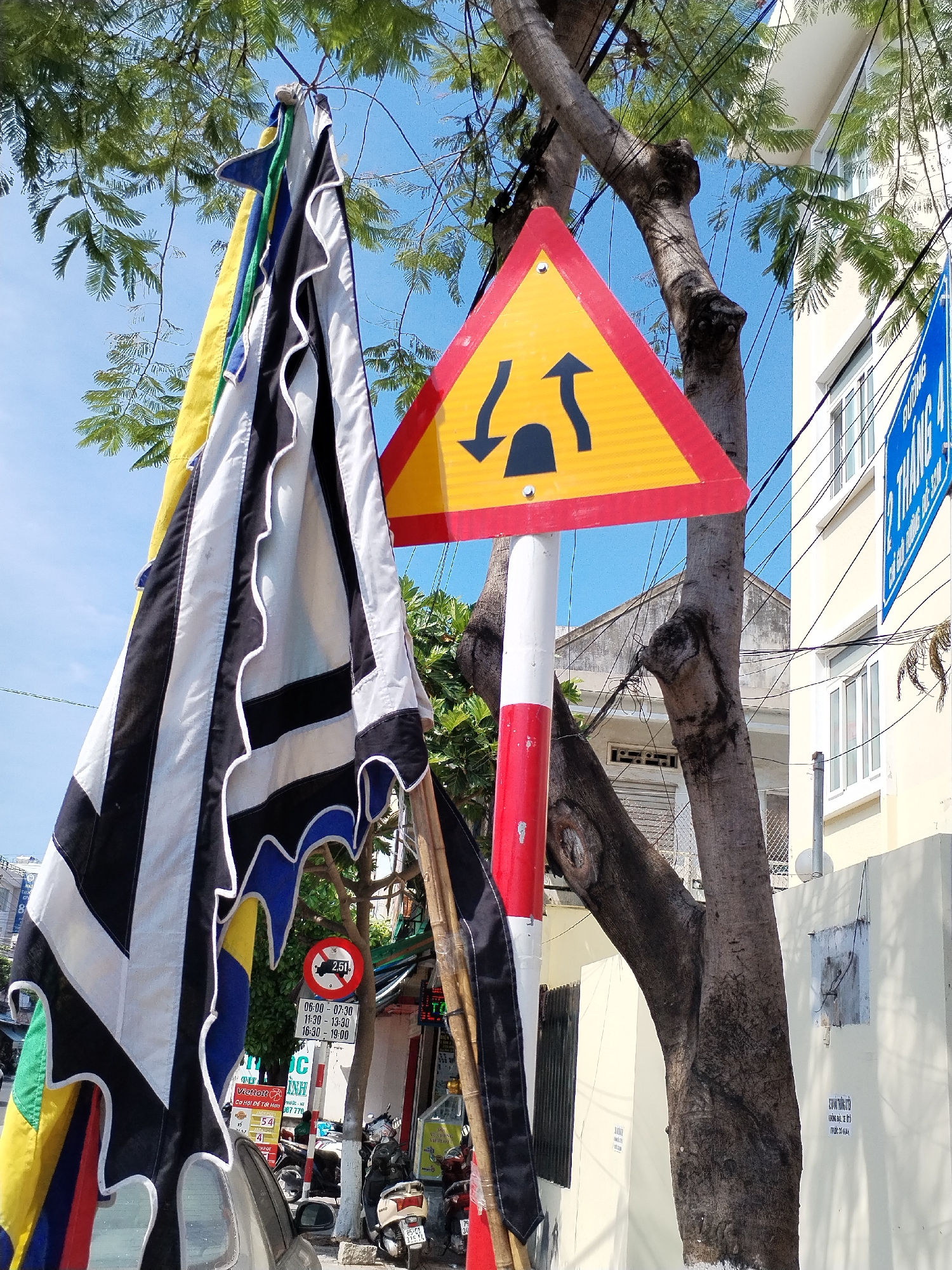 The image depicts a street scene in Bac Nha Trang Ward, Vietnam, on a sunny day. The foreground is dominated by a large, multi-colored flag with black, white, yellow, blue, and green stripes, hanging from a tree. To the right of the flag is a triangular yellow traffic sign with a red border, warning of a two-way traffic ahead. The sign is mounted on a red and white striped pole.
Behind the traffic sign, a large tree trunk splits into two main branches, its bark textured and weathered. In the background, a light yellow building with multiple windows is visible. Several electrical wires are strung across the scene, supported by poles and trees.
Further in the background, on the right side of the image, a blue sign with white text is mounted on the building. Below the traffic sign, a smaller circular red and white sign with a black symbol and text indicating "2.5t" is visible, along with a schedule of operating hours. A bamboo pole leans against the tree.
The street is lined with parked motorcycles, suggesting a typical urban environment. A portion of a silver car is visible on the far left. The ground is paved, and some vegetation, including trees and leafy branches, is present throughout the scene. The sky is clear and blue.