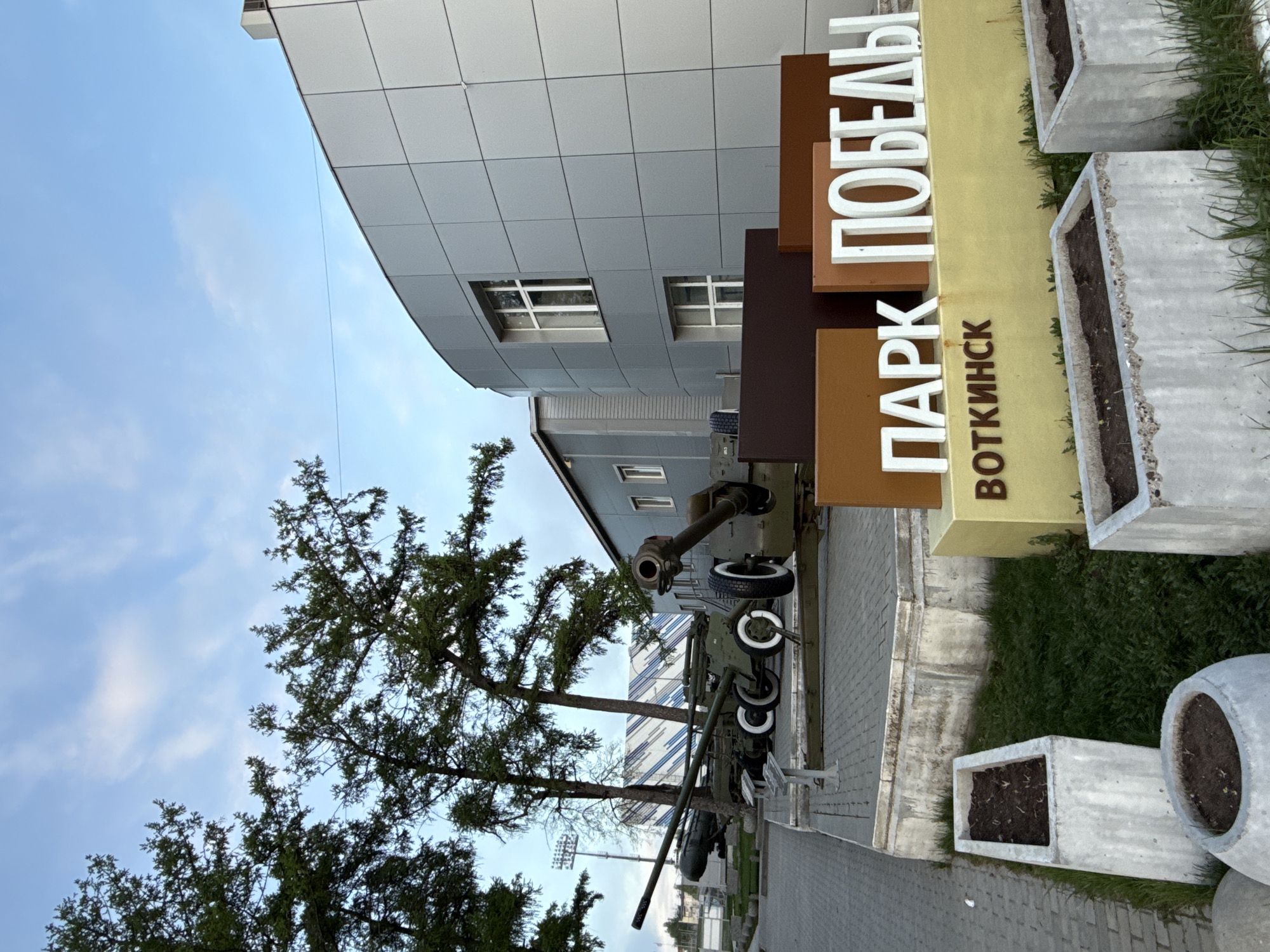 The image depicts a monument in Votkinsk, Russia, near the "Park Pobedy" (Victory Park). A Soviet-era anti-aircraft gun is displayed prominently. A modern building with a gray tile exterior serves as a backdrop. A sign reads "Парк Воткинск ПОБЕДЫ" in white letters on a brown and yellow background, which means "Park Votkinsk Victory." Flower planters are visible along the sidewalk. A tree stands to the left of the anti-aircraft gun. The sky is light blue with some clouds.