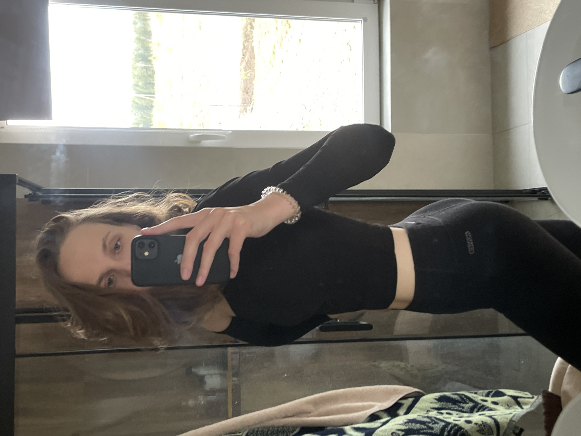 A young woman is captured in a mirror selfie, appearing to be indoors in a well-lit room, possibly a bathroom or a space with a large mirror and window. She has long, light brown hair and is wearing a form-fitting black long-sleeved cropped top and black leggings. A silver-toned beaded bracelet adorns her right wrist, and she holds a black smartphone, likely an iPhone, in her right hand, positioning it to take the reflection. Her expression is neutral, with her eyes focused on the phone's camera.
The background, visible through the reflection, features a large window that reveals bright outdoor greenery, indicating it is daytime. A faint wisp, possibly smoke or steam, is visible near the upper left side of the reflected window area. Below the window is a light-colored wall, and to the right, a curved white fixture, possibly part of a sink or toilet, is partially seen. In the foreground, below the woman's reflected body, lies a dark, reflective surface, likely a countertop. On this surface, a patterned towel with green, blue, and white designs is visible, alongside a plain light-colored towel.
The brand name "GYMSHARK" is clearly visible on the waistband of her black leggings. The Apple logo is also faintly discernible on the back of the smartphone. The setting is Podshivalovo, Russia.