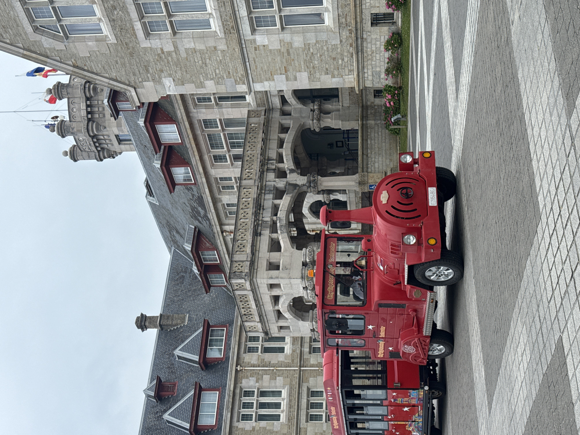 This image depicts the Palacio de la Magdalena in Santander, Spain. In the foreground, a bright red tourist train, resembling a locomotive with passenger cars, is parked on a cobblestone plaza. The train features various decals, including "City Sightseeing Santander." The building behind it is made of light-colored stone and has a grand architectural style with multiple windows, a balcony, and a tower. The sky is overcast. The overall scene suggests a tourist attraction or a public space in Santander.