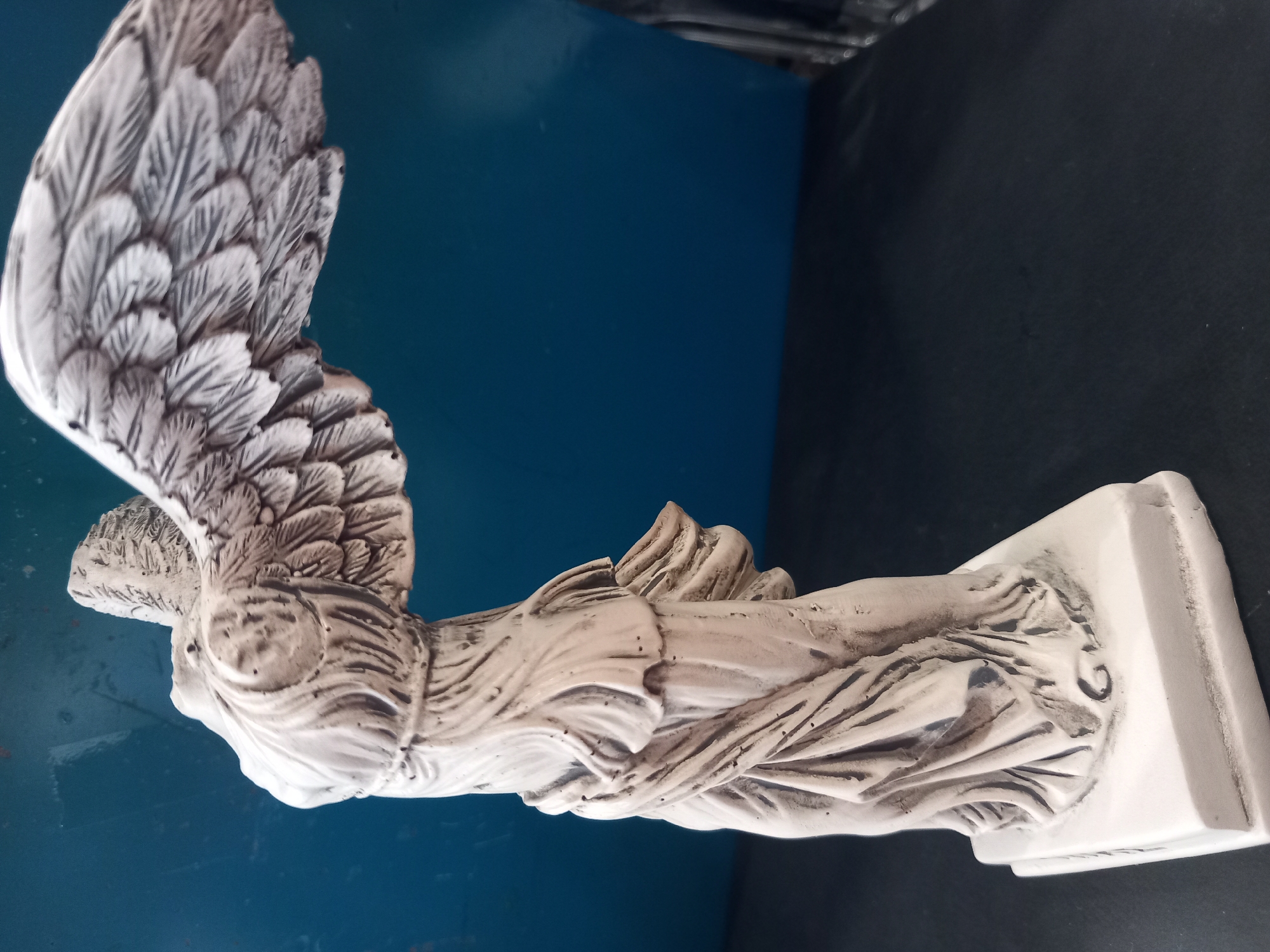 A white statue of Nike, the Greek goddess of victory, stands on a dark surface against a blue background. The statue is depicted with wings outstretched, and her draped clothing appears to be blowing in the wind. The statue is white with some darker shading that highlights the folds of the fabric and the details of the wings. The background is a solid blue. The statue is positioned at an angle, with its right side facing the viewer.