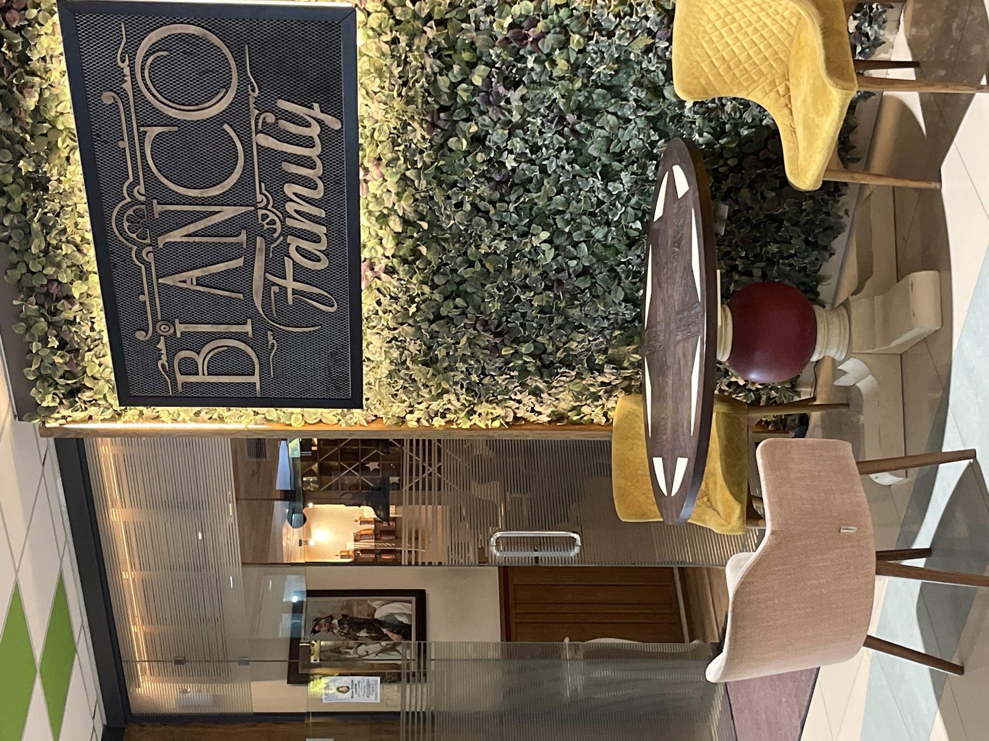 The image shows an outdoor seating area in front of a restaurant. There is a dark wood table with a pedestal base, surrounded by three chairs: one pink, and two yellow. The table and chairs are positioned in front of a wall covered in artificial greenery. A sign that reads "BIANCO Family" is mounted on the greenery wall. To the left, there is a glass door with a metal handle. Behind the glass door, a framed picture of a man on a horse can be seen, as well as shelves with bottles. The floor is tiled. The scene is located in Veliko Tarnovo, Bulgaria.