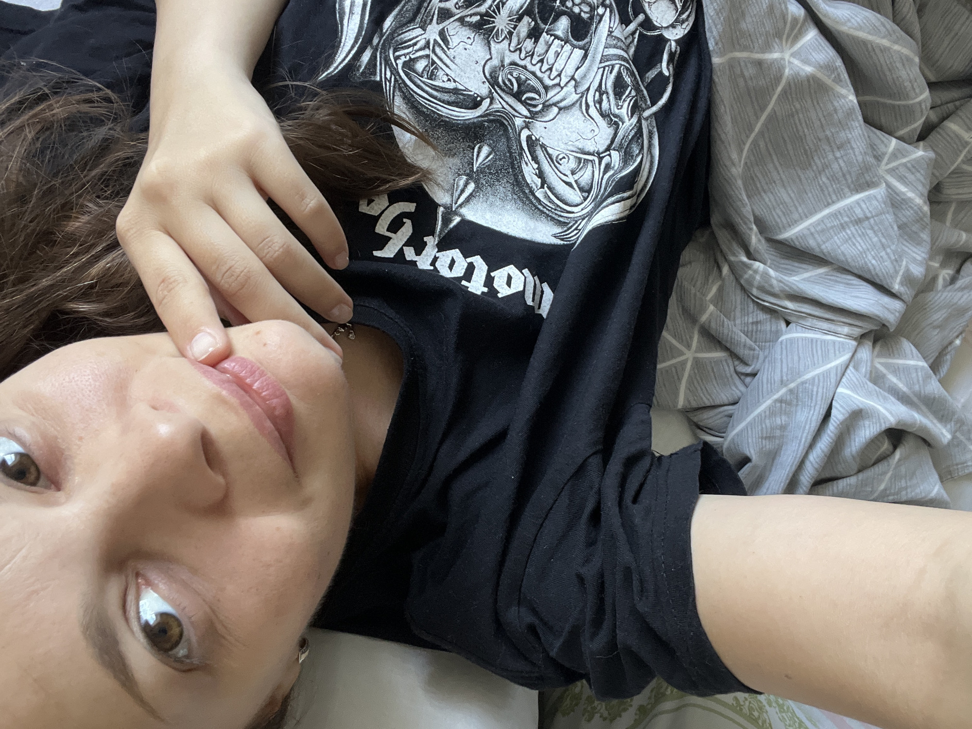A young woman with long brown hair and brown eyes is lying on a bed, looking at the camera. She is wearing a black t-shirt with a graphic print of a skull and the word "motorhead" in white lettering. Her right hand is resting on her chin, and her fingers are touching her lips. The bed is covered with a gray and white striped comforter. The background is out of focus, but it appears to be a bedroom. The image was taken indoors, likely in an apartment or house. There are no specific landmarks or identifying features that would suggest a location in Tolyatti, Russia.