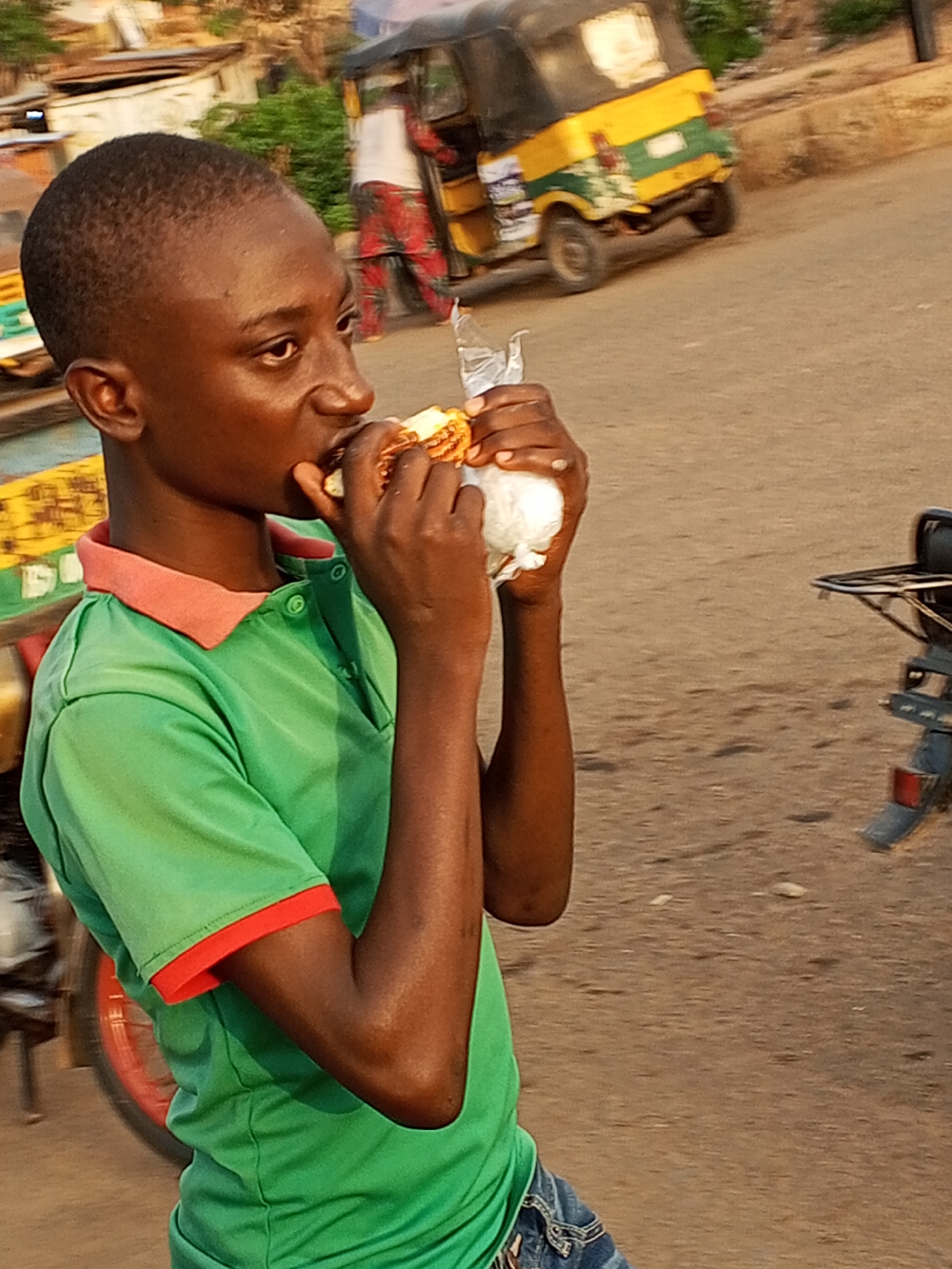 In Gwong, Nigeria, an adolescent male with dark skin and a closely cropped haircut stands near a street, consuming food. He is dressed in a green polo shirt featuring red trim on the collar and sleeves. He uses his right hand to hold a partially eaten piece of food, while his left holds what appears to be white bread in a plastic wrapper. The street environment includes a yellow and green motorized tricycle with a partially visible passenger. A motorcycle is also partially visible in the foreground on the right. The road surface is dirt or asphalt.