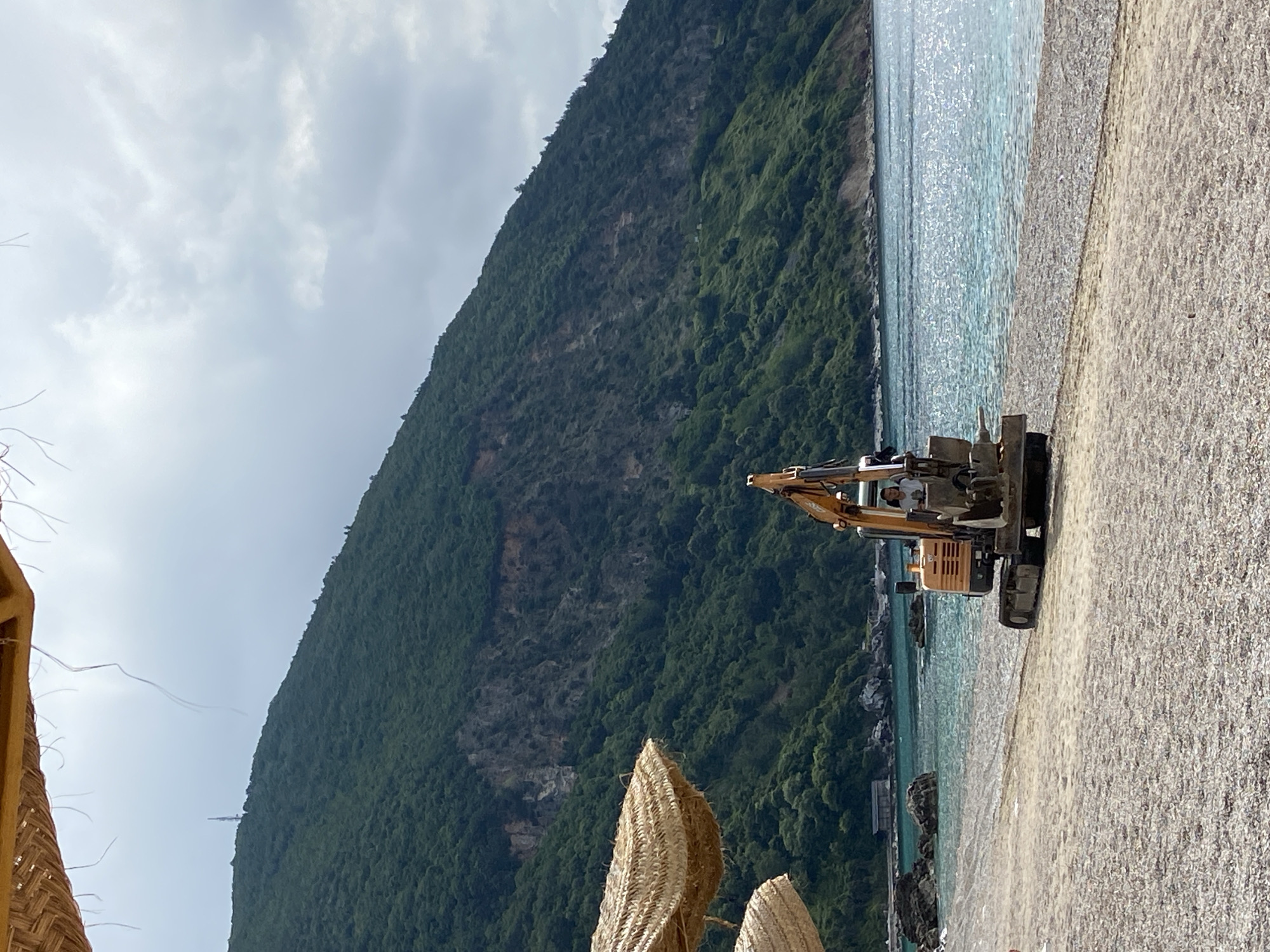 A landscape shot of a beach in Prijevor, Montenegro, shows a mountain covered in lush green vegetation and a light blue sky with white clouds. Turquoise water meets a beach covered in small pebbles. A construction vehicle sits on the beach, facing the water. Part of a woven structure is in the lower left corner, and two smaller woven structures are in the lower center.