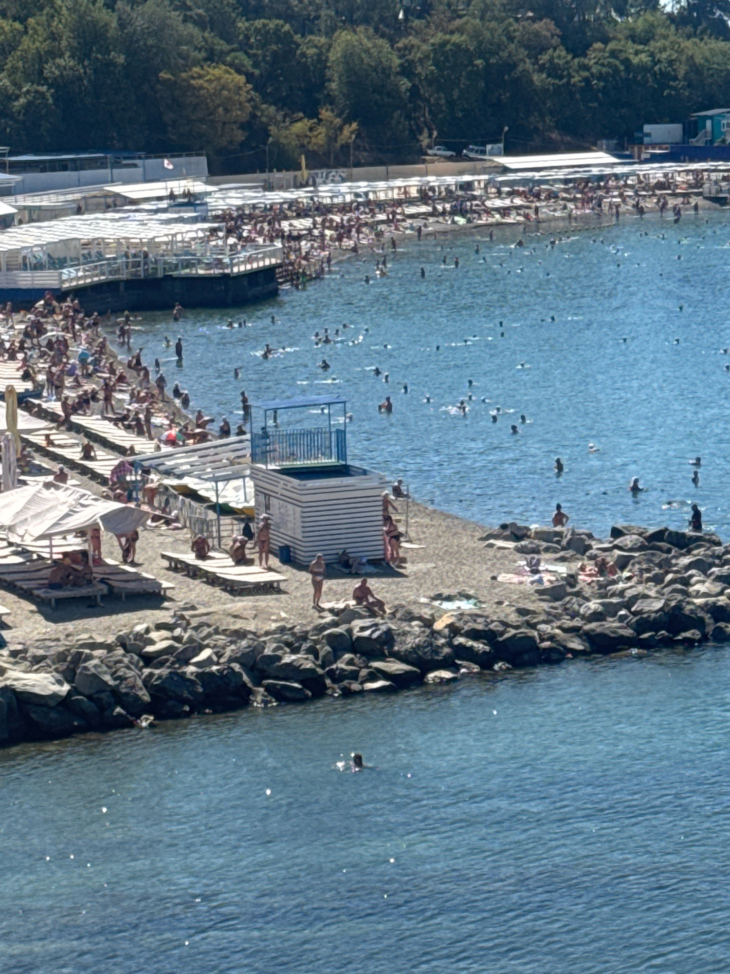 A high-angle, bright, sunlit view of a crowded beach in Anapa, Russia. The scene is dominated by a large number of people enjoying a sunny day at the seaside. In the foreground, a rocky breakwater stretches into the sea, with people sunbathing and sitting on the rocks. To the left of the breakwater, a beach area is covered with lounge chairs and umbrellas, and people are lying on towels or walking. Further back, a pier extends into the water, lined with more umbrellas and loungers, and the beach behind it is densely populated with sunbathers. The sea is a clear blue, with many people swimming and wading. A white and blue lifeguard station with a raised platform is situated on the rocky shore. In the background, a tree-lined embankment with buildings and structures is visible, suggesting the proximity of the city. The image captures a lively and bustling atmosphere of a popular summer resort.