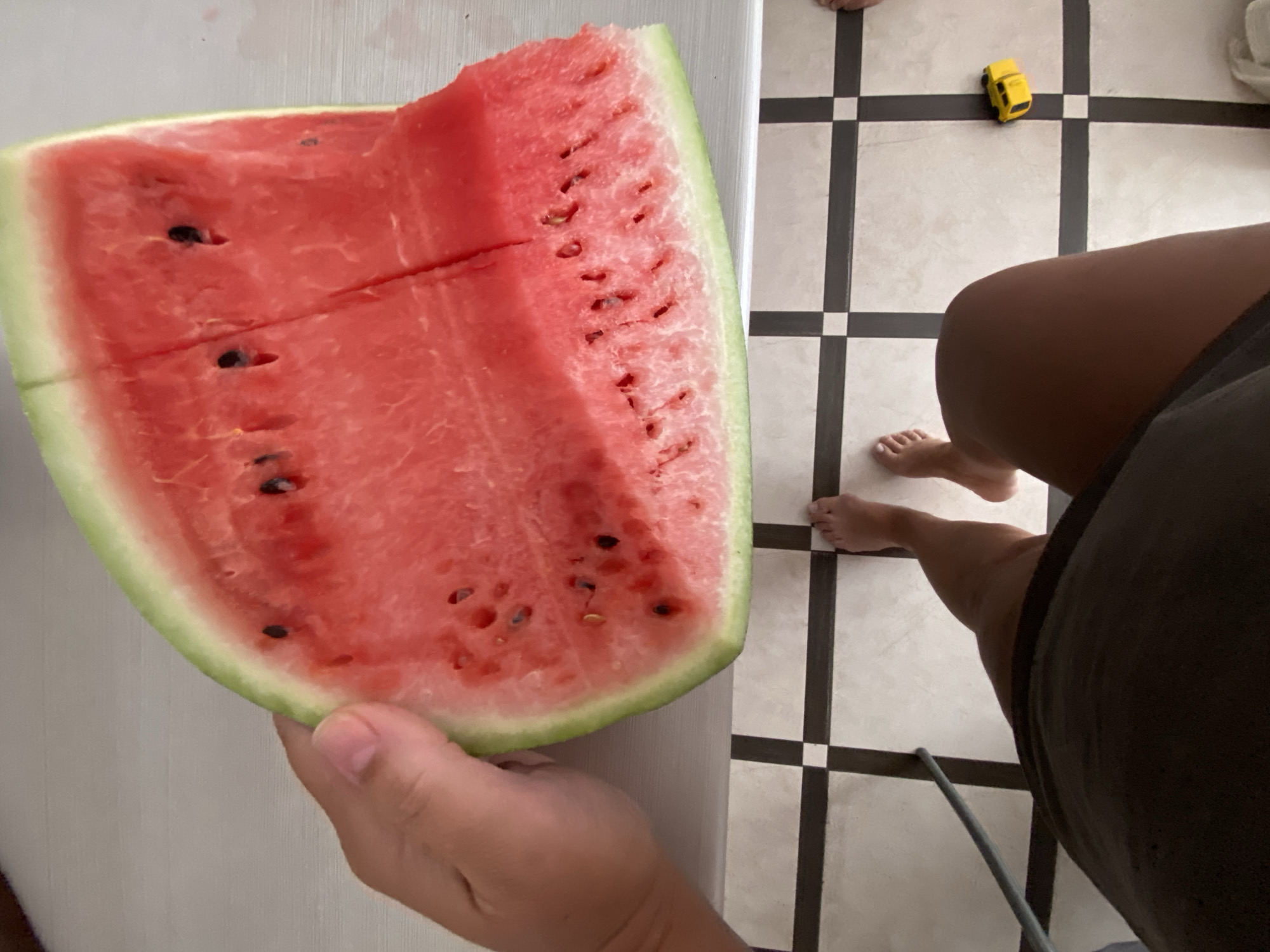 The image is taken from a high-angle perspective, looking down at a person holding a slice of watermelon. The watermelon is a vibrant pink with black seeds and a green rind. The person's hand, which is holding the watermelon, is visible on the left side of the frame. Below the watermelon, a tiled floor with a black and white checkered pattern is visible. Two bare feet are standing on the tiled floor, indicating the person is wearing no shoes. The person is wearing dark-colored shorts or a skirt. To the right of the feet, a small yellow toy car is on the floor. The background appears to be a light-colored surface, possibly a table or countertop. The scene does not contain any specific landmarks or details that would place it within the city of Sochi, Russia.