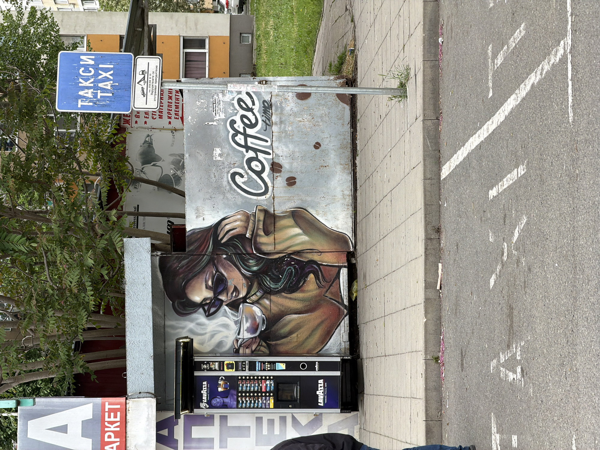 The scene depicts a street in Burgas, Bulgaria, featuring a Lavazza coffee vending machine adorned with a mural of a woman enjoying a cup of coffee and the word "Coffee". Above the mural is the phrase "Coffee Time". Adjacent to the vending machine is a sign indicating a taxi stand and a tow-away zone. A tree is visible to the left of the vending machine, partially obscuring a sign. The sidewalk and a portion of the street with painted markings are also visible. An unknown individual with blue jeans and black clothing is shown from the waist down.