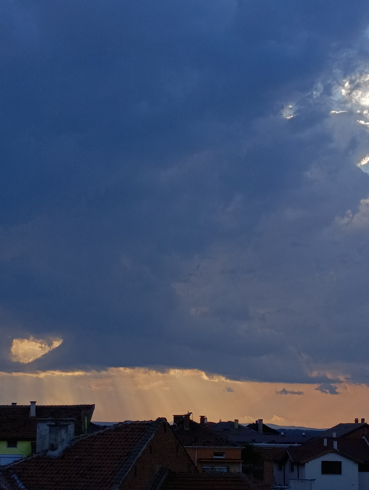 An eye-level shot reveals a sky filled with dark, overcast clouds, partially obscuring the sun. A narrow band of yellowish-orange light filters through the clouds and illuminates the rooftops of numerous houses in the city of Svilengrad, Bulgaria. The buildings consist of both tile and flat rooftops in varying colors and styles. In the background, a distant horizon is barely visible through the haze.
