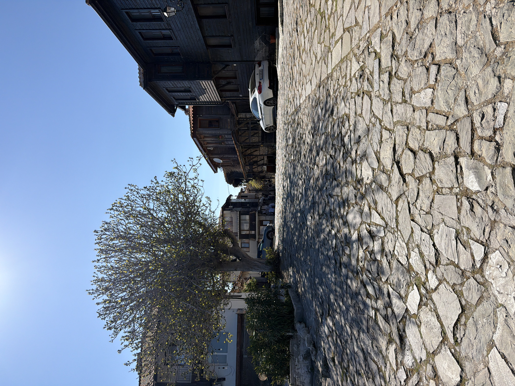 This is an outdoor scene in the city of Nesebar, Bulgaria, featuring cobblestone pavement occupying the right side of the frame. The left side shows buildings, a tree with sparse leaves, and a portion of a blue sky. The buildings are multi-storied with a mix of materials, including dark wood and lighter colored plastered walls. A white car is parked along the street. The sun is visible in the lower left corner. The orientation of the image is rotated counterclockwise.
