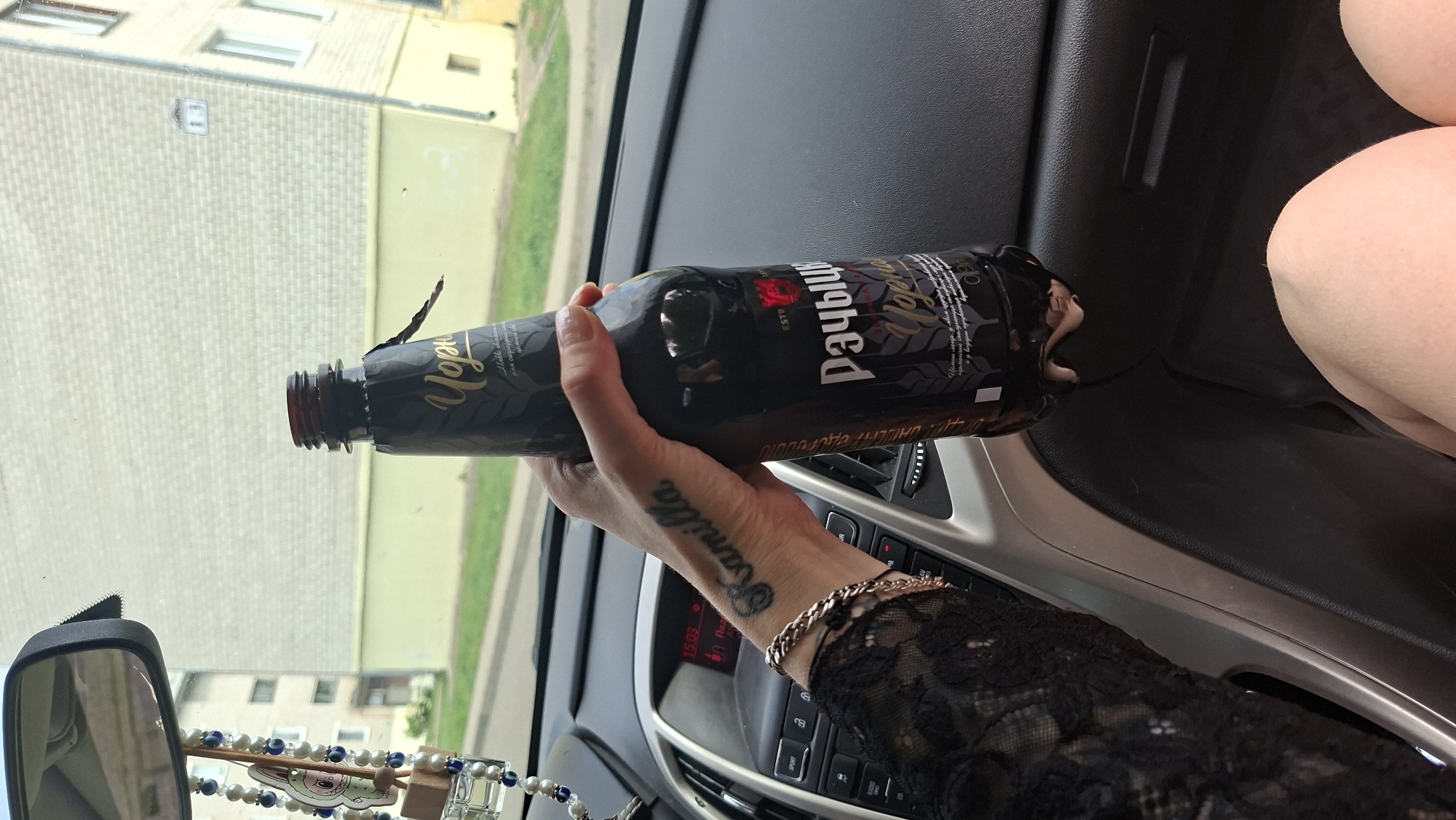 A person's hand, adorned with a bracelet and a tattoo that reads "Kamilla", is holding a dark glass bottle of beer inside a car. The bottle has a label with Cyrillic text, including the brand name "Рапчыца" and "Чорная". The car's dashboard and a rearview mirror are visible in the background, suggesting the car is parked or stopped. Outside the car, a light-colored building with a yellow base is visible, consistent with the architecture found in the city of Myadzyel, Belarus. The overall scene appears to be a casual moment captured during a drive or a stop in Myadzyel.