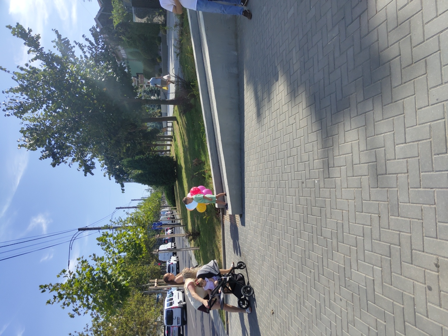 In Chișinău, Moldova, on a sunny day, a mother and her young child are captured on a paved sidewalk adjacent to a road lined with trees. The mother, wearing a white top and dark shorts, is bending down to attend to a baby in a stroller, which is black and has four wheels. Nearby, a young boy, dressed in a light green shirt and shorts, holds a cluster of three balloons—one red, one pink, and one yellow. He stands on the grass beside a low, concrete barrier that separates the sidewalk from a slightly elevated grassy area.
The road features several parked white vans and cars. In the background, more trees and buildings are visible, contributing to a typical urban landscape. The sky is a clear blue with a few wispy clouds. The scene suggests a leisurely moment, possibly a walk in the park or a break during an outing.