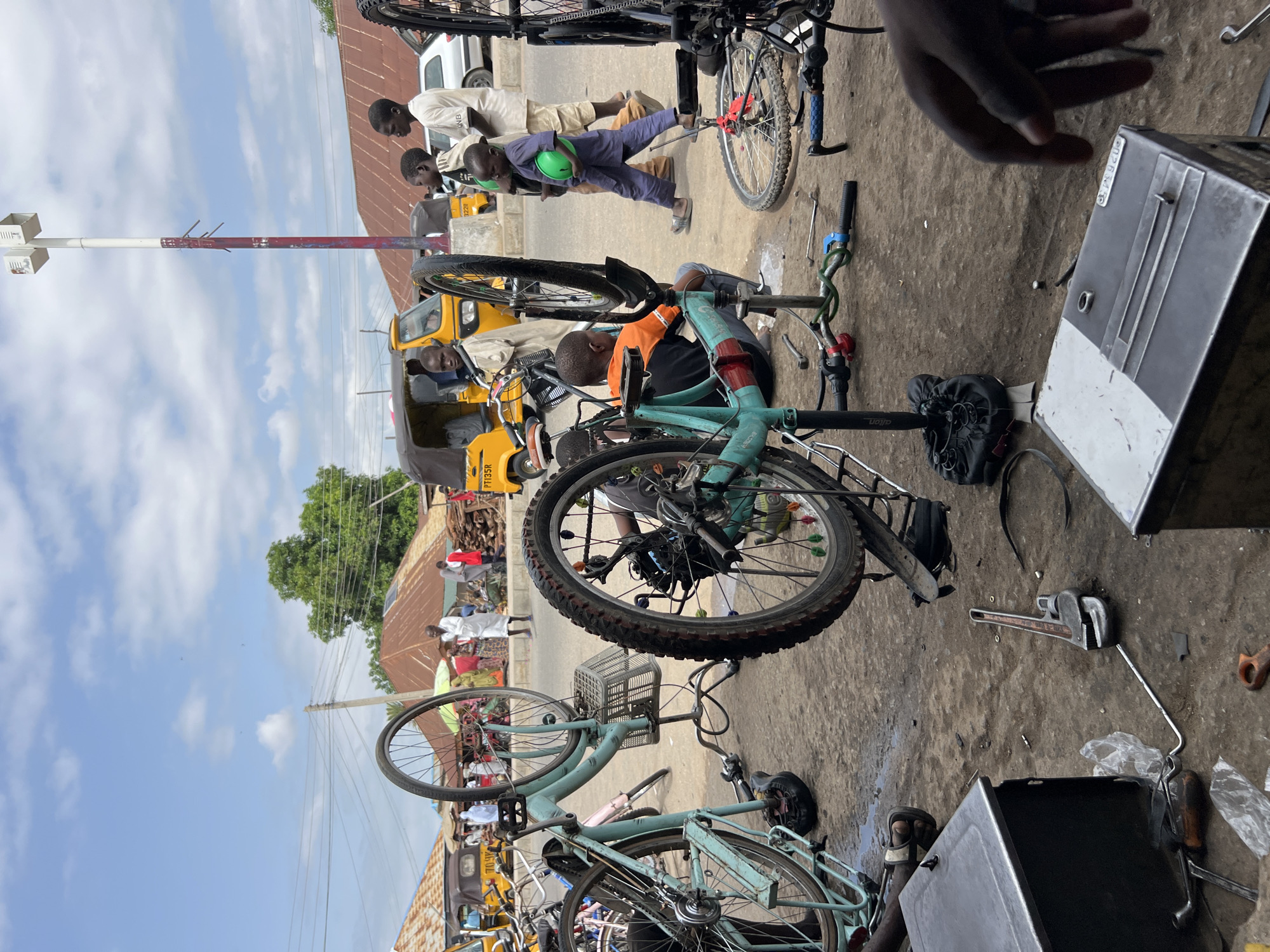 The image depicts an outdoor bicycle repair shop in Jalingo, Nigeria, under a partly cloudy sky. Several bicycles, some disassembled, are the central focus. A turquoise bicycle is prominently displayed on a repair stand, its rear wheel removed. Another disassembled bicycle with a mint green frame is visible to the left. In the foreground, a person's hand is in view, positioned near a metal toolbox. Various tools, including a large adjustable wrench, are scattered on the ground. Several children are present: one in an orange shirt is seated near a bicycle, another child in a blue outfit walks by holding a green helmet, and other children are standing and observing. In the background, yellow auto-rickshaws and other vehicles are parked along a street lined with buildings featuring corrugated metal roofs. Adults are also visible in the background, engaged in activities typical of a marketplace. The ground is paved with a mix of concrete and dirt, with some puddles indicating recent rain. The overall scene conveys a busy street-level commercial activity in an urban Nigerian setting.