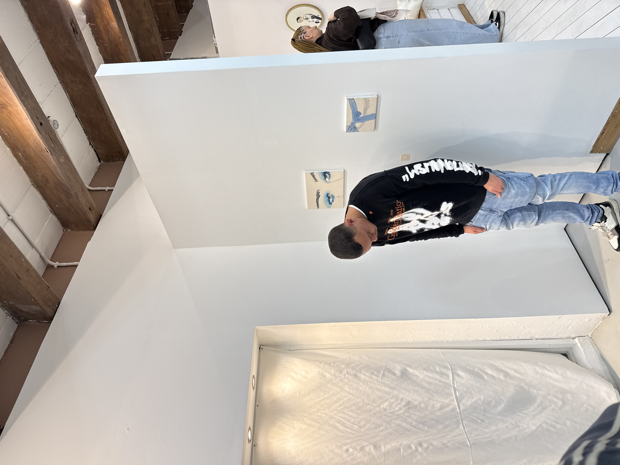 The image depicts an interior space in an unknown city. The scene includes a person wearing a black hoodie and light blue jeans, looking at art on the wall. Another person is present on an elevated platform, wearing a brown shirt and light blue jeans. The room features white walls with exposed wooden beams, suggesting a modern or renovated space. Two pieces of artwork are visible on the wall. Below, a white curtain or covering is evident, potentially obscuring a window or another room element.
