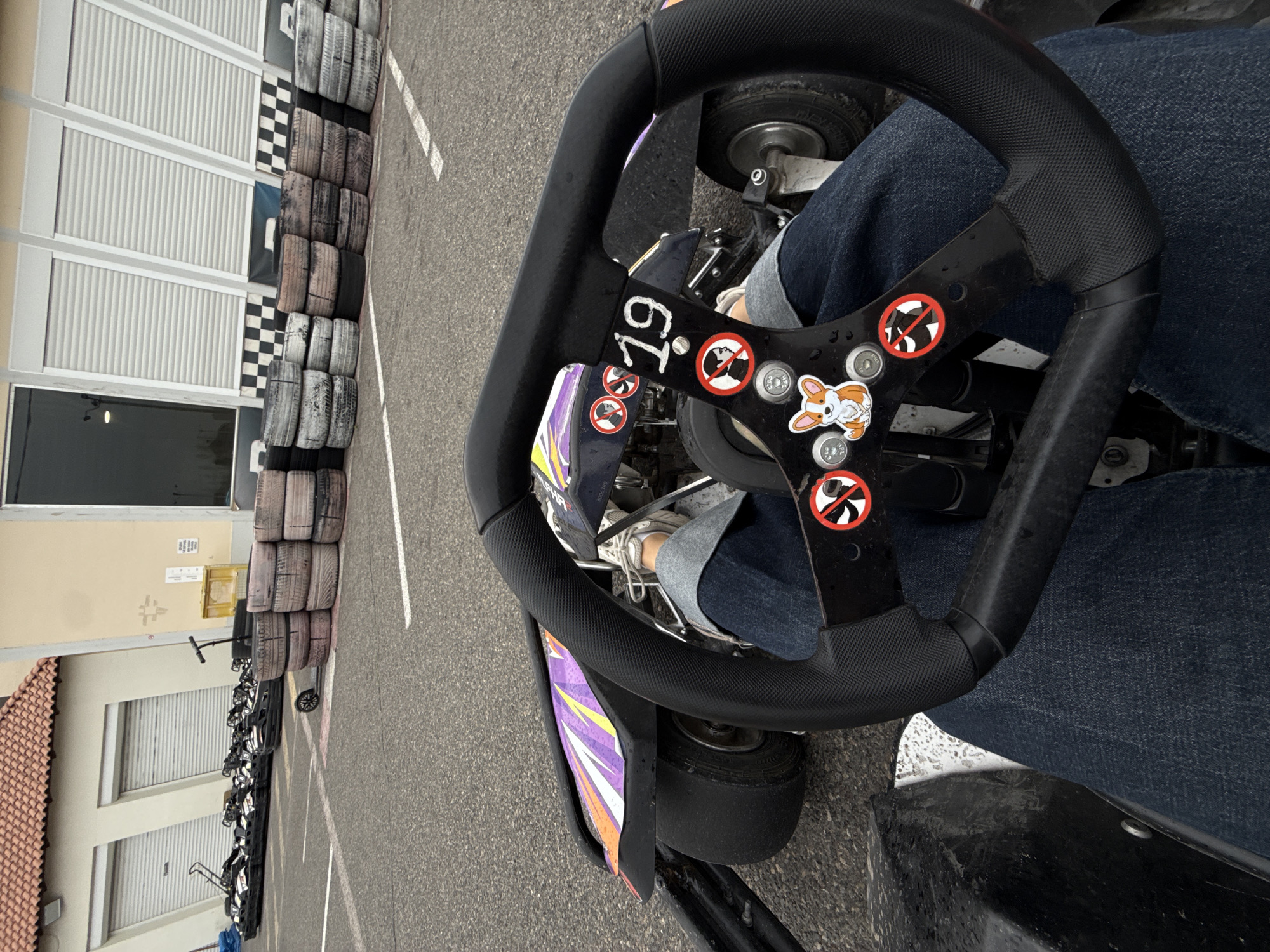 The image captures a first-person perspective from inside a go-kart at what appears to be a racing track in Yerevan, Armenia. The main subject is a person's legs, clad in blue jeans and white sneakers, positioned within the go-kart's cockpit. The black steering wheel dominates the foreground, with visible details including the number "19" in white lettering on its center spoke. Around the "19," there are several red circular stickers with a crossed-out cigarette symbol, indicating "no smoking." A cartoon sticker of a corgi dog is also prominently displayed on the steering wheel's center.
The go-kart itself is partially visible, showing parts of its purple and orange bodywork on the left. The asphalt track surface is gray, with white lines painted on it. To the left, a long line of stacked used tires forms a barrier along the edge of the track, behind which is a building with multiple white roller shutters. Further in the background on the left, more go-karts are lined up, suggesting a queue or storage area. Another building with a light yellow wall and a section of terracotta roof tiles is also visible in the background. The lighting suggests it's daytime, possibly overcast, given the lack of harsh shadows. The image is rotated 90 degrees clockwise.