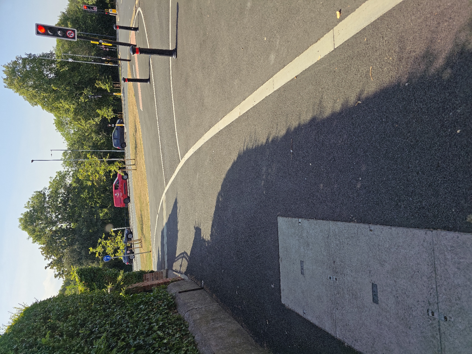 The image depicts a sunny day on a street corner in Stockton-on-Tees, United Kingdom. A lush green hedge lines the left side of the frame, adjacent to a low stone wall and a concrete manhole cover. The road surface is asphalt with clear white lane markings. In the background, several vehicles are visible, including a red van and a dark-colored car. Traffic lights are present, with one showing a red signal. Several black bollards with red tops are positioned along the edge of the road. Trees and streetlights are also scattered throughout the scene, indicating an urban or suburban environment. The overall impression is one of a typical street scene on a clear day.