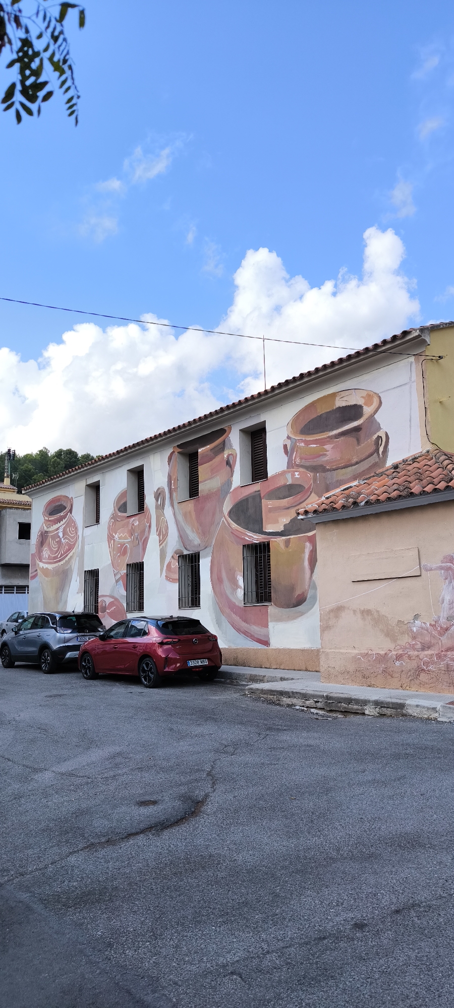 The image shows a mural on the side of a building in Fanzara, Spain, depicting various terracotta-colored amphorae. The mural covers a significant portion of the building's facade, featuring large, stylized pots with intricate patterns. The building itself has white walls and brown-shuttered windows. In front of the building, a paved street is visible, with two cars parked along the curb: a gray SUV and a red hatchback. The sky above is blue with scattered white clouds. The street appears to be in a residential or semi-urban area, with some greenery and other buildings visible in the background. The overall scene suggests a sunny day in a Spanish town.