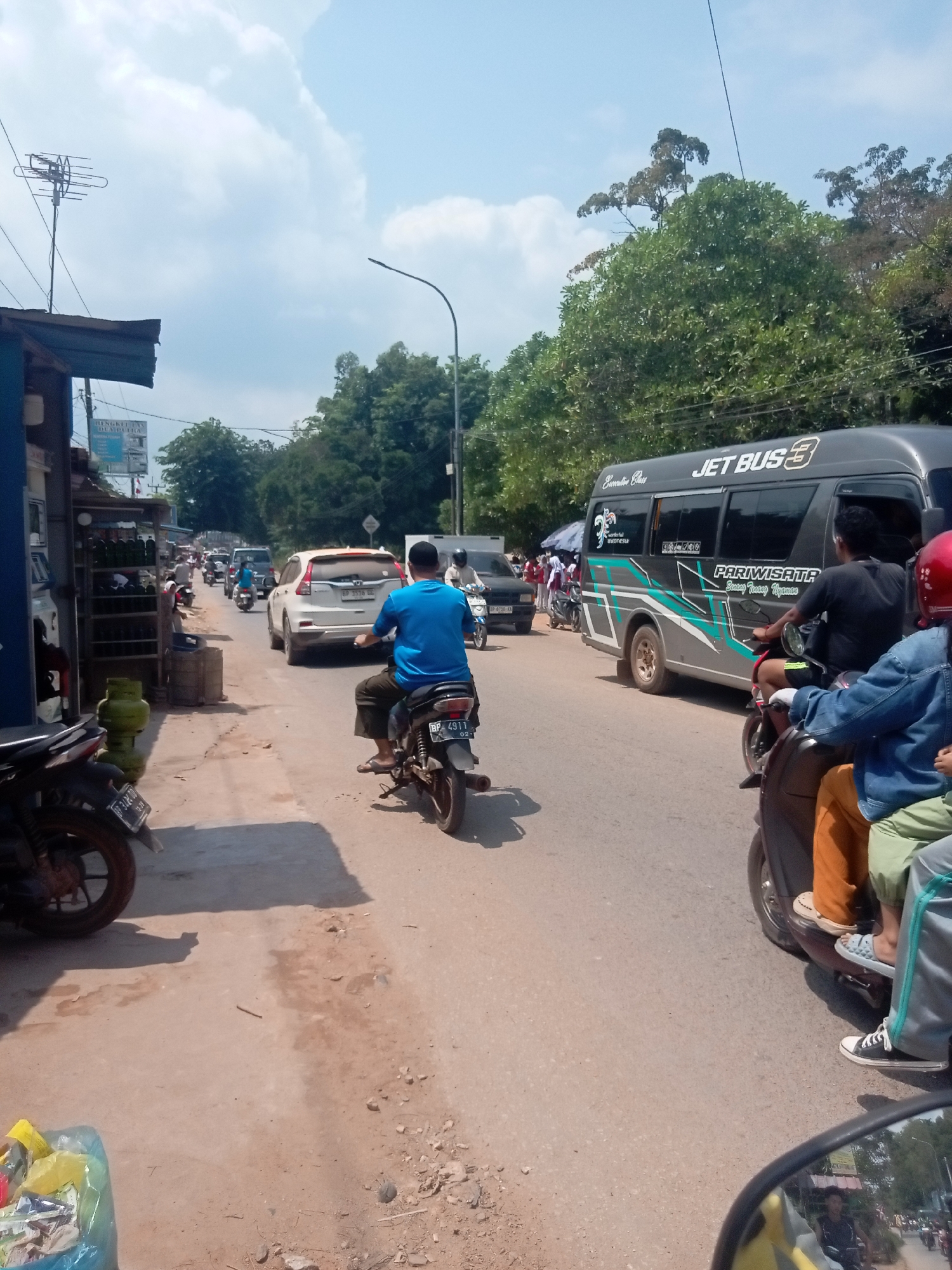 The image depicts a street scene in Batam, Indonesia, under a partly cloudy sky. The road appears to be a two-lane street with moderate traffic. On the right side of the road, a dark gray minivan with "JET BUS 3" and "PARIWISATA" branding is driving. The minivan has green and teal graphics. Behind the minivan, a person wearing a red helmet on a scooter is visible, with another person behind them wearing a blue denim jacket and orange pants.
On the left side of the road, there's a sidewalk with various objects, including shelves filled with bottles, a blue gas tank, and a blue trash bag filled with items. Several motorcycles are parked or moving along the street. A white SUV is in the middle of the road, and further down, other vehicles and motorcycles are present. A person wearing a blue shirt and dark pants is riding a motorcycle in the foreground, with their back to the camera. Trees line the right side of the street, and some structures, possibly shops or stalls, are visible on the left. An antenna is mounted on a pole on the left side of the image. The overall impression is a busy urban street with everyday activity.
