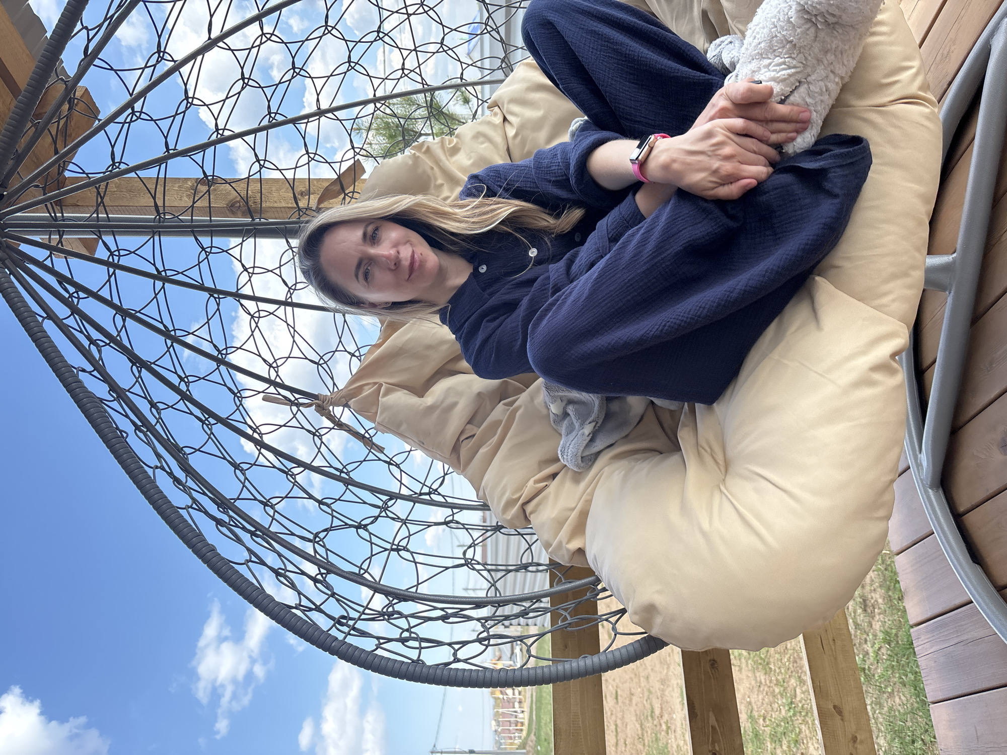 A woman is seated in a gray metal, egg-shaped hanging chair with beige cushions, resting her hands on a light gray item in her lap. She wears a dark blue shirt and pants and a pink smartwatch. The chair is situated on a wooden deck or porch. Outside the chair, there is a view of a grassy area, some buildings, and a blue sky with scattered clouds. Based on the provided location, this scene likely depicts a personal leisure space in or near Chaikovsky, Russia.