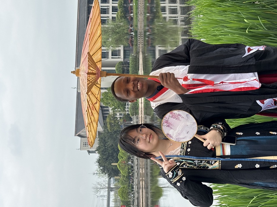 The image displays two young adults, an East Asian female and a Black male, posing outdoors in traditional Chinese attire in Jiangjin, China, on an overcast day. The female, positioned on the left, wears spectacles and a black long-sleeved robe featuring elaborate gold, blue, and white embroidery of floral and nature motifs, layered over a white undergarment and a teal pleated skirt. She holds a round, long-handled fan with a purple floral design in her left hand and makes a peace sign with her right hand; a dark-colored smartphone is visible tucked into her sash. The male, on the right, wears a black traditional Chinese robe with red and white trim, featuring a red decorative Chinese knot with tassels. He holds a large, open yellow-orange oil-paper umbrella with a wooden handle in his right hand. Both individuals are smiling broadly. In the immediate foreground, tall green reeds are present. Behind the subjects, a calm body of water reflects the sky and the distant surroundings. The background features modern multi-story buildings with some traditional architectural elements, fronted by manicured landscaping with small trees and bushes along the water's edge. Further in the distance on the far left, power lines and a bridge structure are visible against the light gray, cloudy sky.