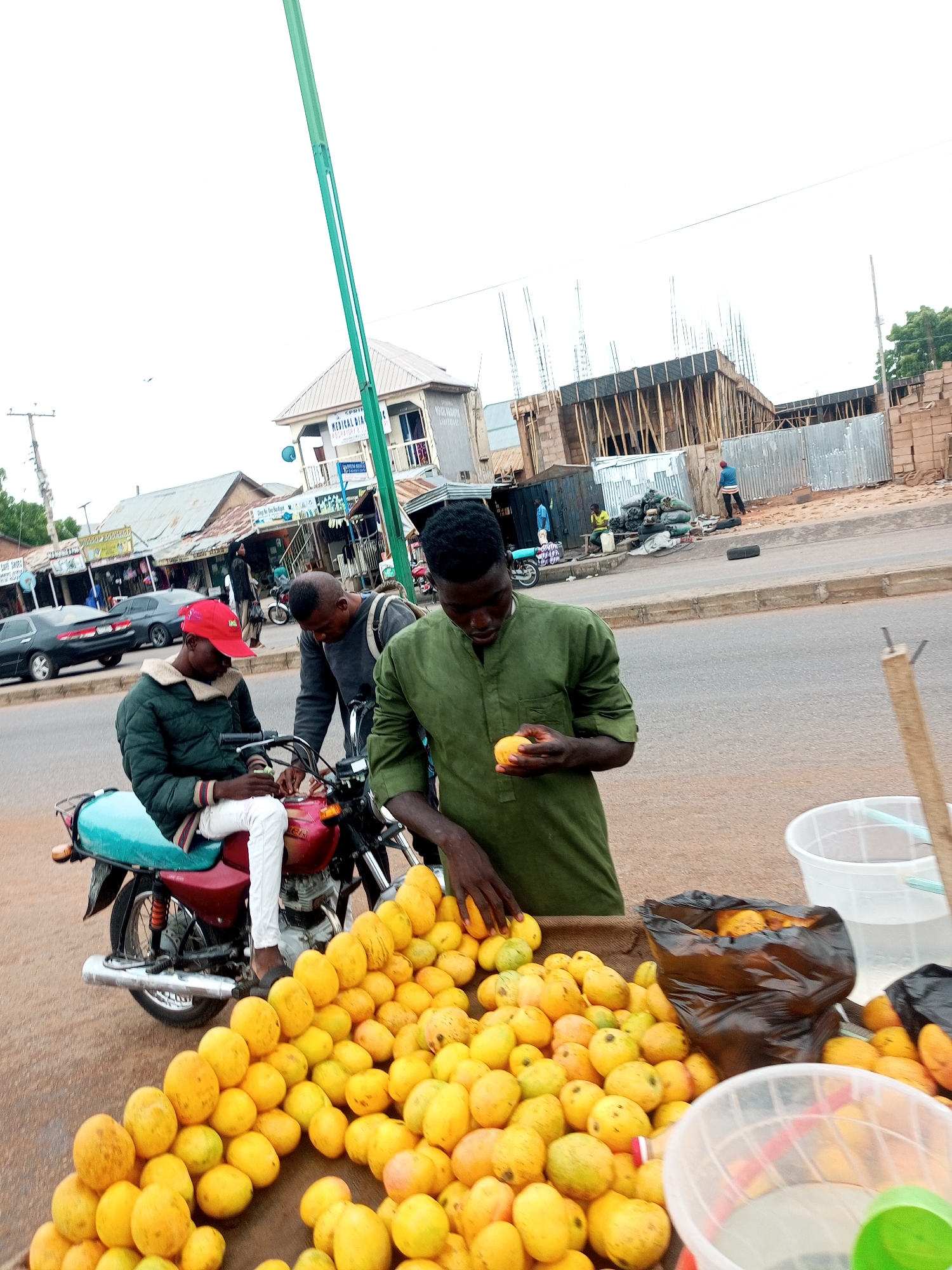 A medium shot captures a street scene in Tempure, Nigeria, with an emphasis on a fruit stand and people interacting around motorcycles. In the foreground, a large pile of ripe mangoes is displayed on a table. A man in a green garment is holding a mango, seemingly in the process of selling or preparing it. Beside the mangoes, a black plastic bag bulges with more fruit, and a clear plastic bucket filled with liquid is visible. To the left, a motorcycle with a turquoise seat is parked, and a man in a green jacket and red cap sits on it, looking down at his hands. Another man in a grey sweatshirt stands next to him, also looking towards the motorcycle. In the background, a street is lined with buildings and shops, with cars parked along the side. Construction is underway on a building further down the street, indicated by scaffolding and bare concrete structures. A tall, green pole, likely a street light or signage, extends upwards into the overcast sky. The ground is paved with a reddish-brown material, suggesting it's an outdoor marketplace or roadside vendor area.