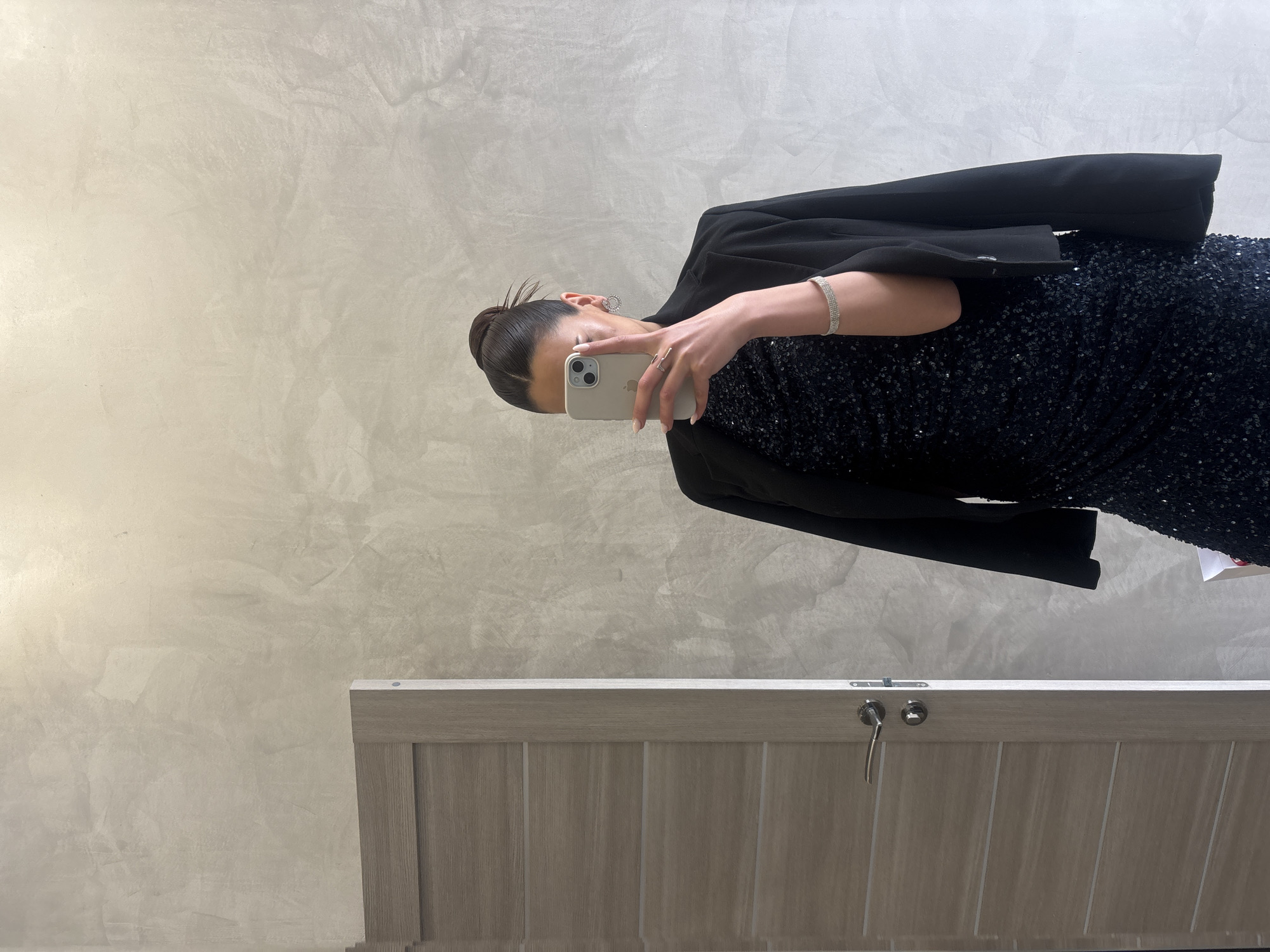 A woman is taking a selfie in front of a mirror. She is wearing a black, sequined dress with a black blazer draped over her shoulders. She has her hair pulled back in a bun and is wearing a bracelet on her left wrist. The background is a light gray wall. The bottom of a wooden door is also visible. The location is Yerevan, Armenia.