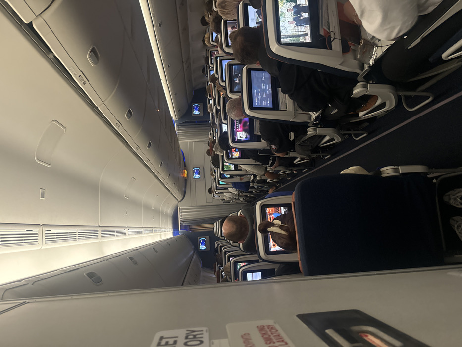 The image captures the interior of an airplane cabin, rotated 90 degrees clockwise. When viewed correctly, it shows a long airplane cabin with passengers seated, likely during a flight.
On the left side of the corrected image, the pristine white ceiling and overhead compartments of the aircraft are visible. The overhead bins are closed, featuring a sleek, modern design with subtle handles and ventilation grates along their lower edge. Recessed lighting strips run along the ceiling and beneath the bins, casting a soft, ambient glow.
On the right side, multiple rows of airplane seats stretch into the distance. Most passengers are seen from behind, settled into their seats. The seats are upholstered in a dark blue or black fabric. Each seatback is equipped with an individual in-flight entertainment (IFE) screen. Many passengers are actively engaged with these screens, which display various content, including what appears to be movies, TV shows, or flight information interfaces. Some screens show people in a wooded scene, while others display menu options. The screens have a light-colored bezel.
In the mid-ground, a passenger with a bald head is visible, looking at their screen, which appears to be showing a brightly colored animation or cartoon. Further back, another passenger is wearing a dark hoodie, and brown shoes are visible beneath their seat. The general activity is passive entertainment, typical of a long-haul flight.
The aisle runs down the center of the visible cabin section, separating the rows of seats from the overhead bins and providing a pathway through the aircraft. The floor is covered with a dark carpet.
The overall lighting in the cabin is dim, suggesting that it might be a nighttime flight or that the cabin lights have been lowered for passenger comfort. Small blue accent lights are visible further down the aisle in the distance, possibly marking exits or other facilities.
At the bottom of the original image (which would be in the foreground, near the floor, if corrected), partially visible text is present on what appears to be a sign or a safety card. The legible text includes "...ET TORY" and, below it, "NO SMOKING LAVATORY". The visible part of the sign appears to be white with black lettering.