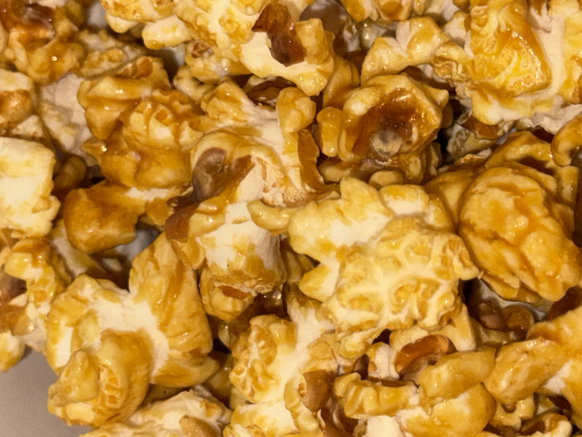 This is a close-up, overhead shot of a pile of caramel popcorn. The popcorn pieces are varied in size and shape, with a glossy, golden-brown caramel coating that glistens under the light. Some pieces show the fluffy, white interior of the popped corn, while others are more densely packed and appear to contain nuts. The texture of the popcorn looks crisp and slightly sticky due to the caramel. The lighting suggests it might be indoors, with the light source coming from above, creating highlights and shadows that give the popcorn a three-dimensional appearance. There is no visible text in the image, nor any indications of a specific time of day, weather conditions, or location context beyond the general context provided. The overall impression is one of a delicious, tempting snack.