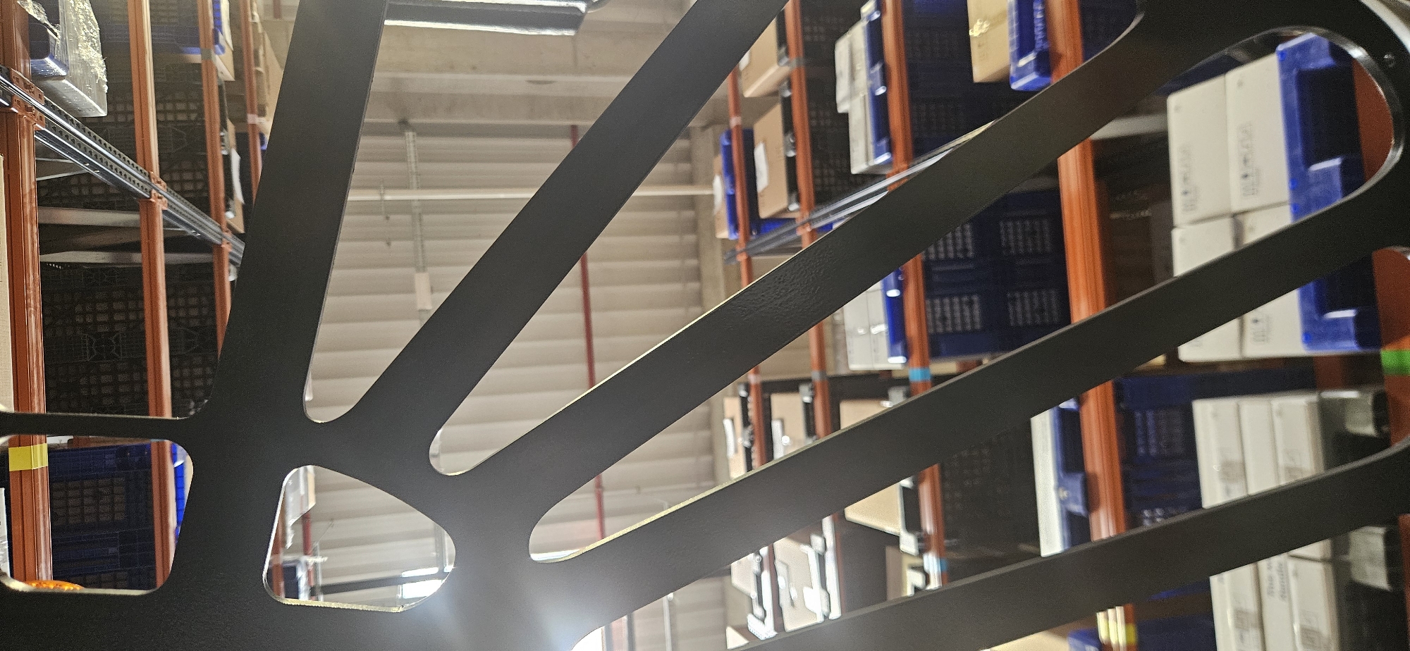 The image depicts a section of what appears to be a large storage warehouse, likely in Plovdiv, Bulgaria. The foreground is dominated by a dark, intricate metal structure with repeating geometric shapes and radiating linear elements. The background consists of rows of metal shelving, filled with boxes and plastic containers, mostly in shades of white and blue. The lighting appears to be predominantly artificial, with bright spots. The scene is still and there are no immediately discernible people present.