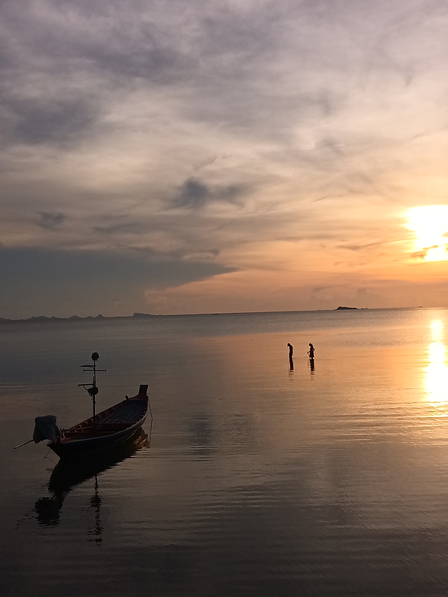 The image depicts a sunset scene in Baan Wok Tum, Thailand. A small, wooden fishing boat with an outboard motor is anchored in shallow water in the lower left of the frame. Two figures, likely people, are standing in the water further out, near the center of the frame. The sky is filled with clouds, with the sun setting on the right side, creating a golden reflection on the water's surface. The horizon line is visible in the distance, with a hint of land. The overall tone of the image is calm and serene.