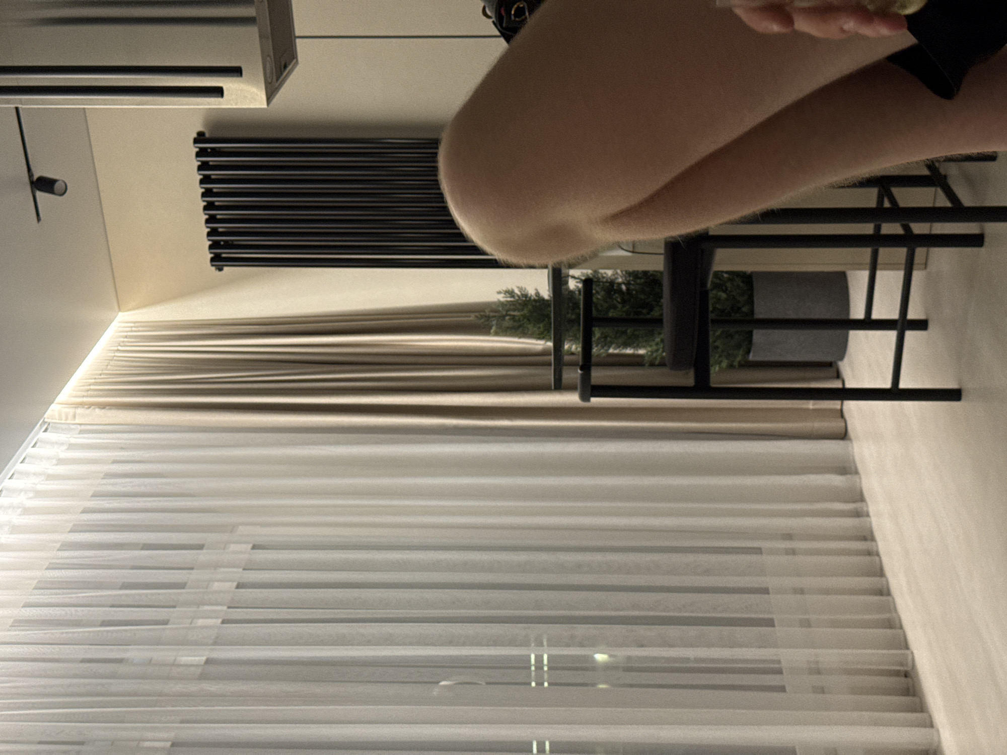 The image depicts an indoor scene, likely a modern apartment or living space. In the foreground, a person's leg is visible, with a close-up on the knee and upper thigh area. The person appears to be relaxed, possibly seated. The background features a sheer white curtain covering a window, allowing diffused light to enter. To the right of the curtain, a set of light brown drapes is visible. Further back, a black radiator is mounted on a light-colored wall, and above it, a track light fixture is installed on the ceiling. A tall, slender black bar stool with a dark grey plant pot on the seat is positioned in the middle ground, with a small, conical green plant inside the pot. The floor is a light-colored wood or laminate. The overall ambiance suggests a contemporary and minimalist interior design. There is no discernible information or visual cue within the image that would indicate the specific location as Izhevsk, Russia.