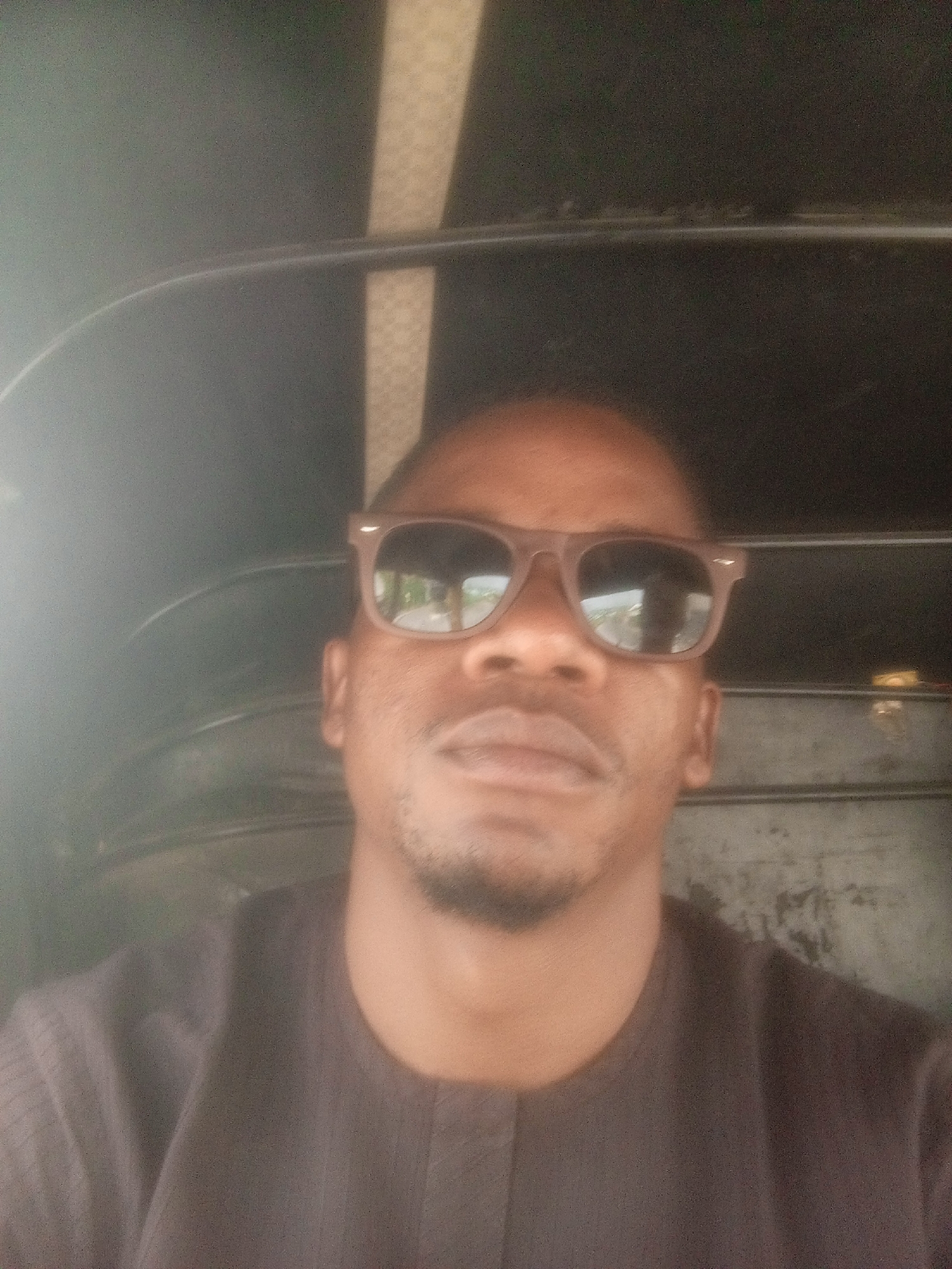 A person is in a commercial tricycle (Keke Napep) with dark glasses on. The person is located in Gwarinpa, Nigeria, based on prior knowledge of the individual. The surroundings inside the vehicle are metallic gray, indicating the interior of the tricycle. The person is wearing a brown shirt.
