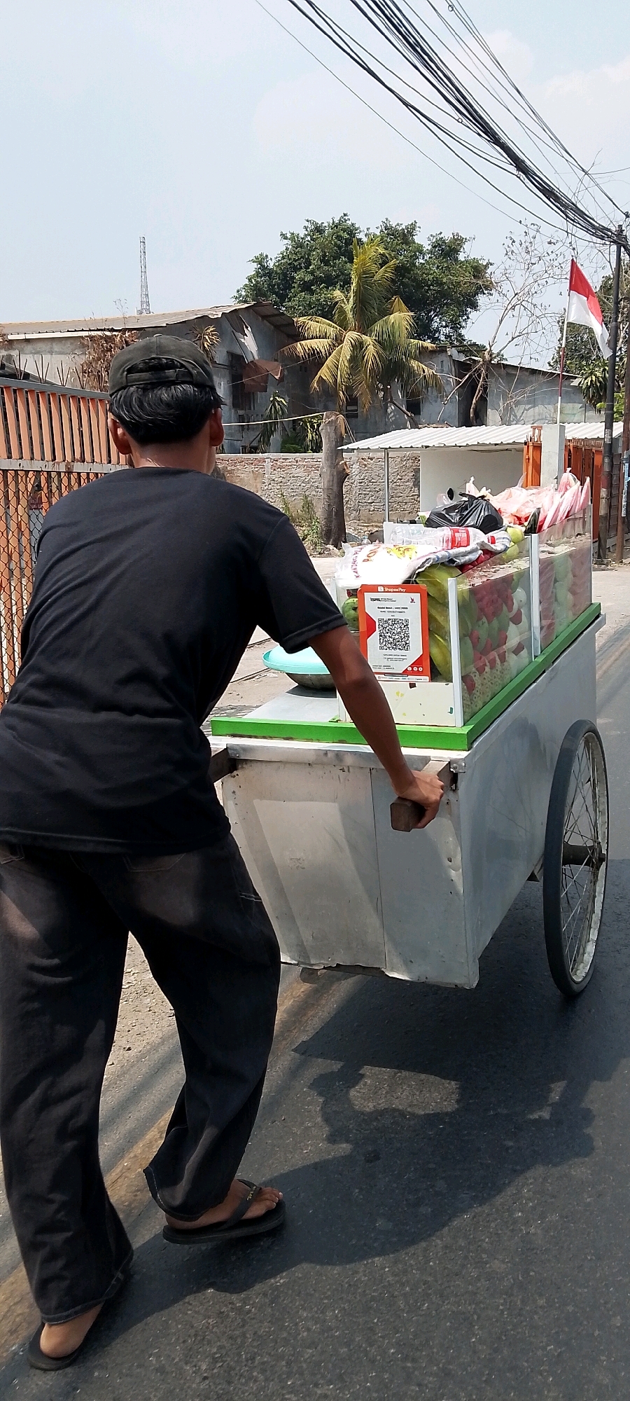 In Bekasi, Indonesia, a person wearing a dark t-shirt, dark pants, and flip-flops pushes a street food cart along a paved road. The cart has a white and green metal body and is filled with various colorful items, possibly fruits or candies, displayed in a clear plastic container. A QR code for ShopeePay is prominently displayed on the front of the cart. In the background, a tall radio tower is visible against a clear sky, alongside trees, a building with a corrugated metal roof, and a white building with a small awning. A small Indonesian flag flies from a pole near the white building. Power lines stretch across the upper portion of the image. The scene is illuminated by sunlight, casting shadows on the road.