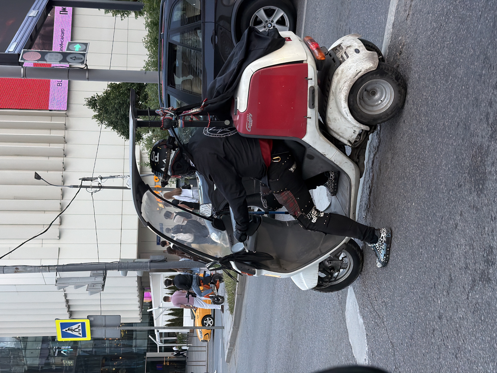 A sideways, medium shot depicts an individual on a three-wheeled vehicle in a city setting. The person is wearing a helmet, dark jacket, and pants with patterned accents. The vehicle is white with a red panel and a partial canopy. It is parked on a street adjacent to a curb. The background includes buildings with varying facades, traffic signs, a traffic light, other vehicles, and pedestrians.