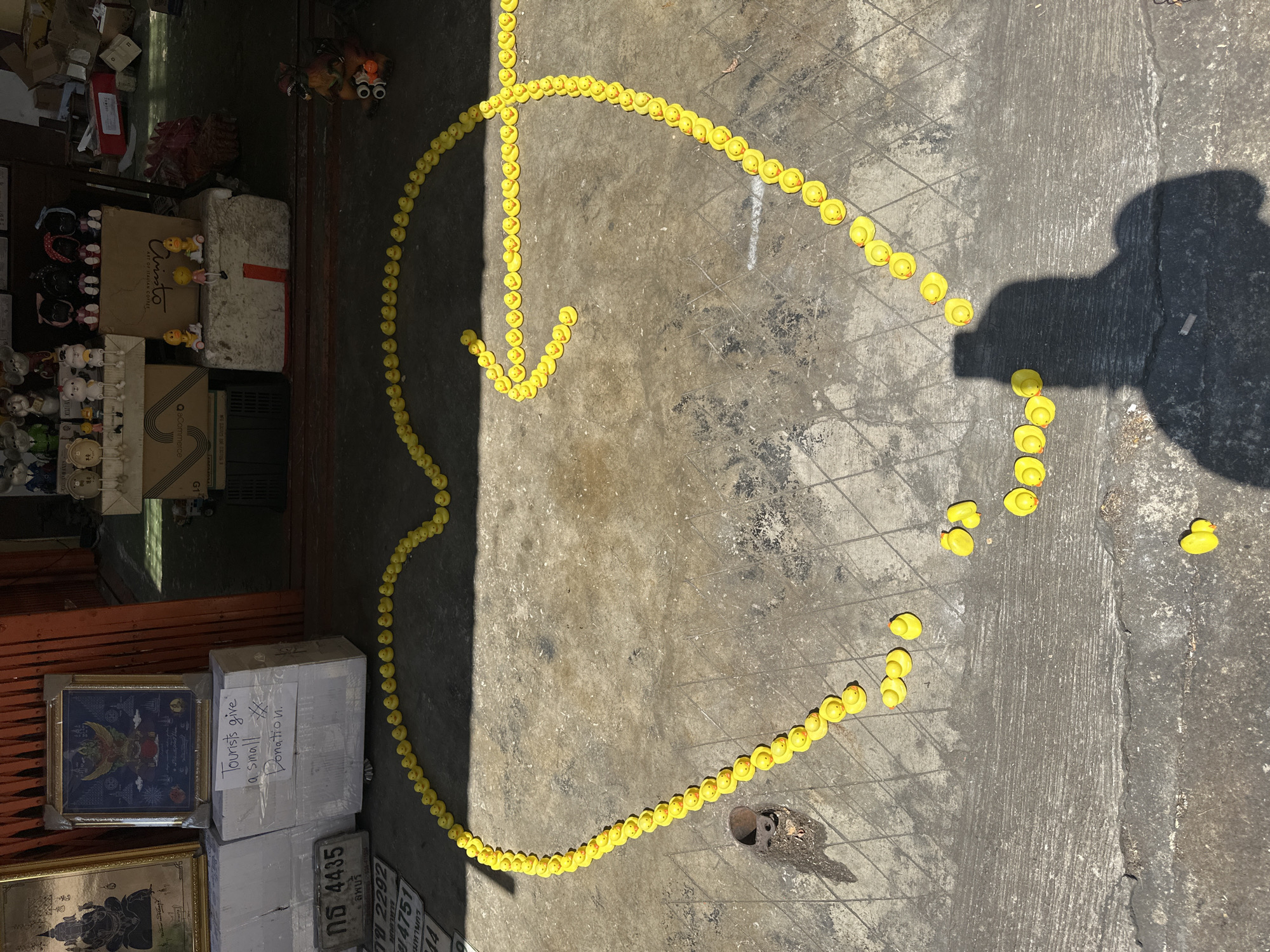 The image shows a collection of small, yellow rubber ducks arranged in the shape of a heart with an arrow extending from one side, lying on a concrete surface. The ducks are arranged in a line, forming the outline of the heart and arrow. To the left of the ducks are several boxes and frames, some containing religious images. One box has the text "Tourists give a small Donation." written on it. There are also license plates and merchandise in the frame. A shadow of a person is visible to the right. The photograph appears to be taken outdoors, likely in a commercial or tourist area in Bangkok, Thailand, given the text on the donation box.