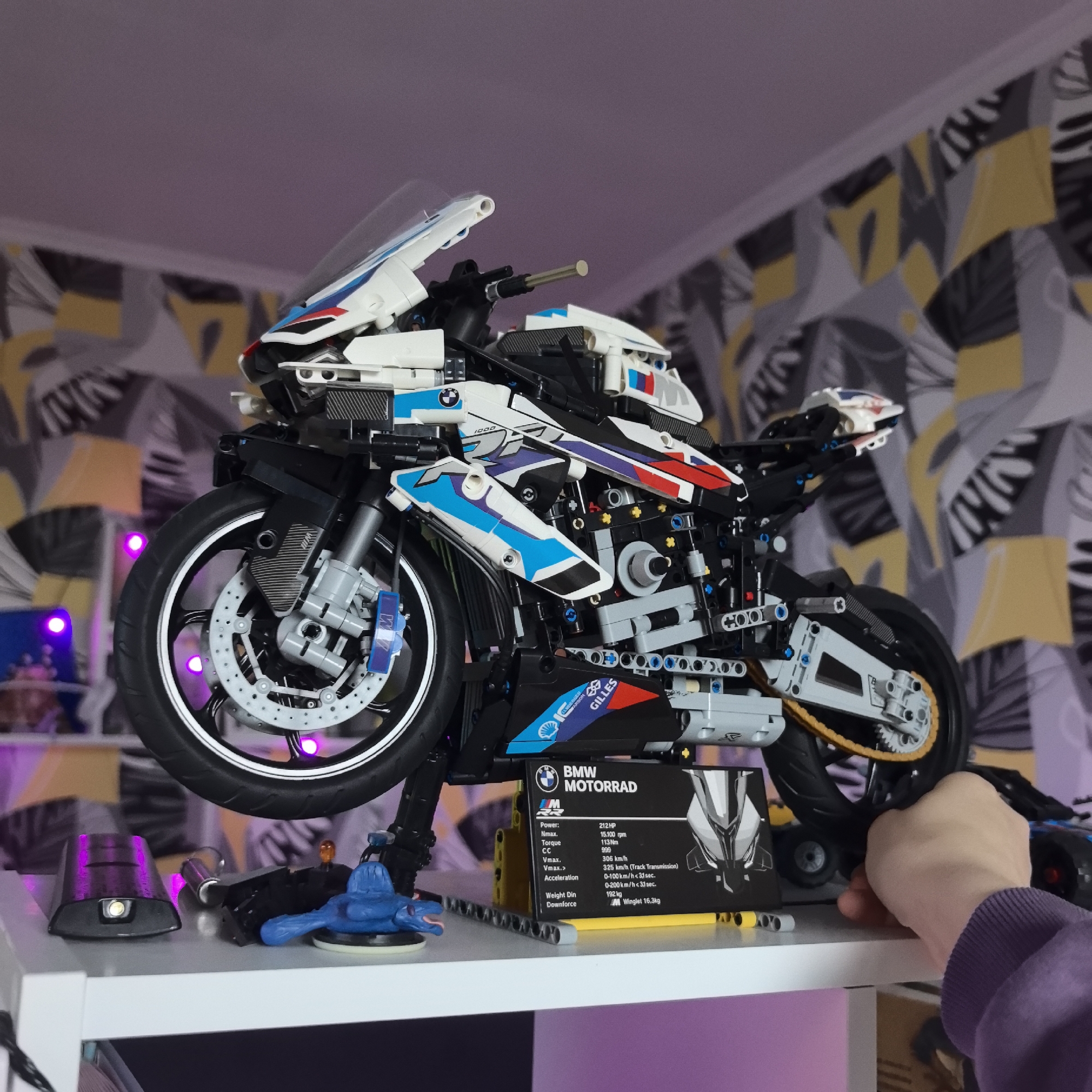 An indoor shot taken in Khimki, Russia, features a highly detailed LEGO Technic model of a BMW Motorrad motorcycle. The model, predominantly white with blue, red, and black accents, rests on a white shelf. Its front wheel is elevated by a stand, giving the impression of motion or display. A person's hand, wearing a purple sleeve, is seen holding the rear of the motorcycle, possibly steadying it or adjusting its position.
The motorcycle model showcases intricate details, including visible engine components, a chain drive, front disc brakes, and fairings adorned with "1000" and "BMW" logos, as well as M-sport branding. Below the motorcycle, on the white shelf, are a few small objects, including a dark light fixture emitting a white glow, a metallic cylindrical object, and a small blue figure.
A prominent black display plaque for the "BMW MOTORRAD" model is positioned on the shelf, listing specifications such as:
* Power: 212HP
* Nmax.: 15,100 rpm
* Torque: 113 Nm
* CC: 999
* Vmax.: 306 km/h
* Vmax. (Track Transmission): 325 km/h
* Acceleration (0-100 km/h): < 3.1 sec.
* Acceleration (0-200 km/h): < 3.3 sec.
* Weight Din: 192 kg
* Downforce (M Winglet): 16.3kg
The background features a patterned wall with abstract shapes in yellow, black, and grey/white. A soft pink and purple glow emanates from behind the motorcycle, likely from decorative LED lighting, adding ambiance to the display. The lighting suggests it is an indoor setting, possibly during evening hours.