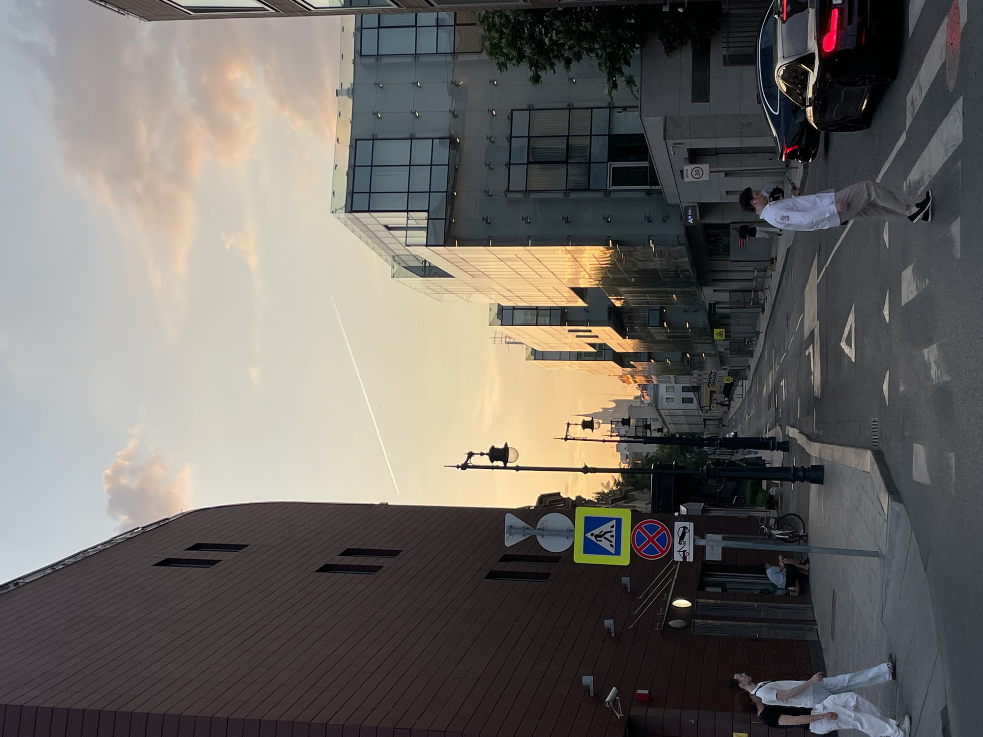 The image shows a street scene in an urban environment. The photograph appears to have been taken at ground level with the camera tilted.
Several buildings are visible. On the left is a multi-story structure with a dark brown facade and small rectangular windows. Adjacent to this building is a modern glass-fronted building reflecting the sunset.
Street lighting is visible along the side of the road, with a lamppost in the foreground near a pole with several road signs affixed. The signs include a pedestrian crossing sign, a no-parking sign, and a sign indicating a cycle path.
On the right side of the street, a person wearing light-colored clothing is crossing a zebra crossing in front of a parked black car. Further down the street, near the bottom edge of the image, two individuals are standing next to the brown building.
The sky is visible in the upper left of the frame, displaying a blend of light blue and orange hues, indicative of either sunrise or sunset. A contrail from an airplane is also present in the sky.
A bicyclist is parked down the street.