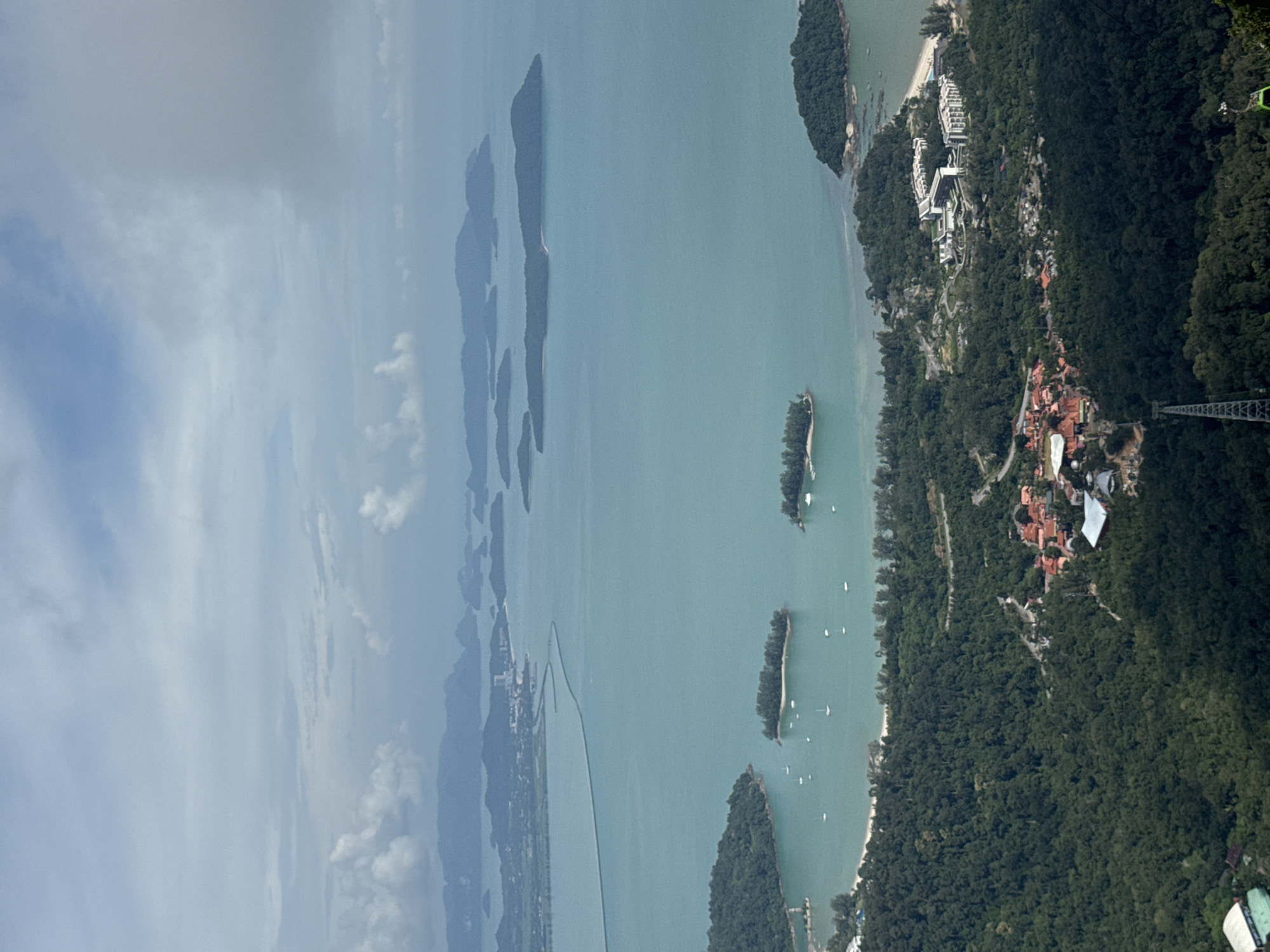 This image is a high-angle view of Langkawi, Malaysia, likely taken from the top of the Langkawi Cable Car. The foreground is dominated by a dense, lush green forest covering the slopes of a mountain. A section of the cable car track, a tall metal pylon and a green cable car cabin, is visible in the lower right portion of the image.
Further down the mountain, nestled within the trees, is a complex of buildings with orange-tiled roofs, appearing to be a village or resort. To the right of this complex, a larger modern resort with white buildings and multiple wings is situated on a coastline with a sandy beach.
The middle ground features a bay with calm, turquoise water. Several small, tree-covered islands are scattered within the bay. Small white sailboats are dotted across the water, indicating a tranquil marine environment. A long, thin bridge or causeway extends across the water in the distance, connecting to a mainland area with more distant, hazy islands.
The background consists of a vast expanse of the ocean with numerous islands of varying sizes, some with visible vegetation and others appearing as darker, mountainous shapes. The sky above is a mix of cloudy and clear patches, with soft, diffused light. The overall impression is of a serene tropical landscape with a blend of natural beauty and developed infrastructure.