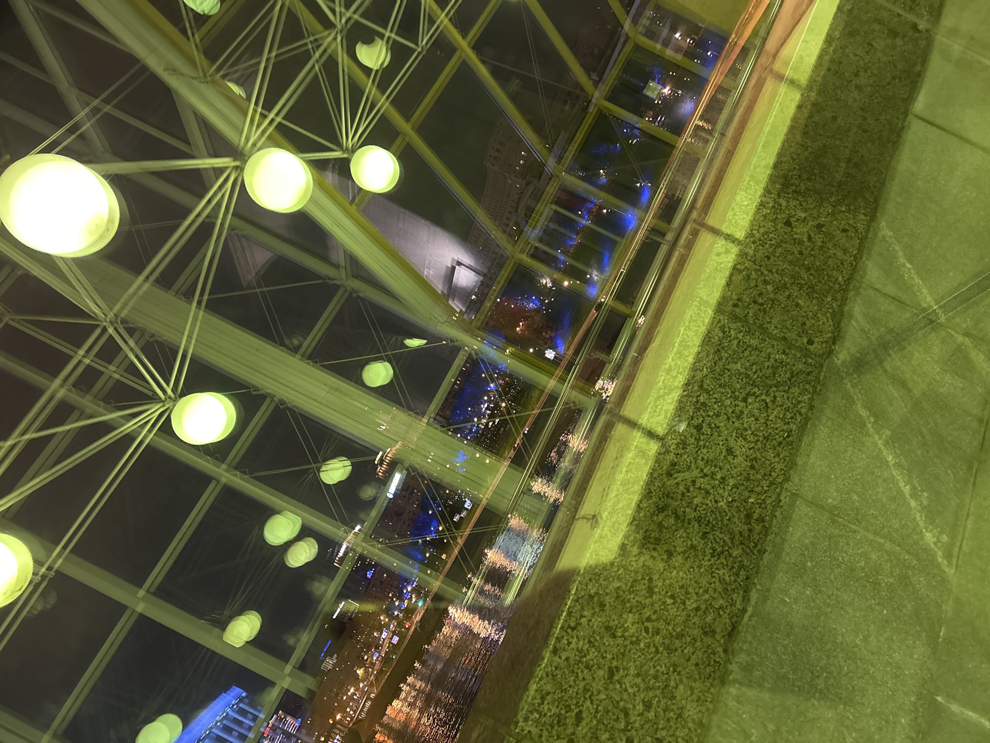 The image shows an interior view looking down through a glass structure with a metal frame, with lights hanging from the frame. The glass provides a distorted reflection of the city lights below, suggesting a tall building. A green, textured surface appears to be the edge of the viewing platform or wall. The photograph is taken at an angle, giving a tilted perspective.