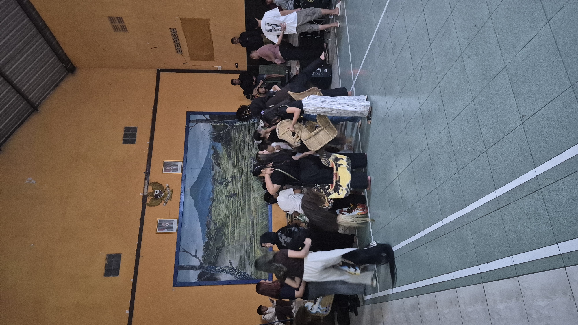 The image depicts an indoor gathering in what appears to be a community hall or gymnasium located in Sumowono, Indonesia. The hall has a high, corrugated metal roof and orange-painted walls. A large mural depicting a mountainous landscape with terraced fields covers a significant portion of the back wall, beneath which hang two framed portraits and a national emblem.
In the foreground, a group of individuals, predominantly young adults, are engaged in various activities. Several people are wearing black clothing, suggesting a cultural or ceremonial context. One person appears to be in costume, with elaborate black hair or a headdress extending to the floor. Others are holding woven baskets or carrying items. Some individuals are standing and observing, while others are interacting closely, possibly preparing for an event or participating in a performance.
To the right side of the scene, a musical setup is visible, including drum kit components and speakers, indicating the presence of a band or musical accompaniment. A few individuals are standing near the musical equipment, dressed casually.
The floor is tiled in a checkerboard pattern, with white lines marking out a court or designated areas. The overall impression is that of a lively, communal event taking place in a functional, multi-purpose space within Sumowono.