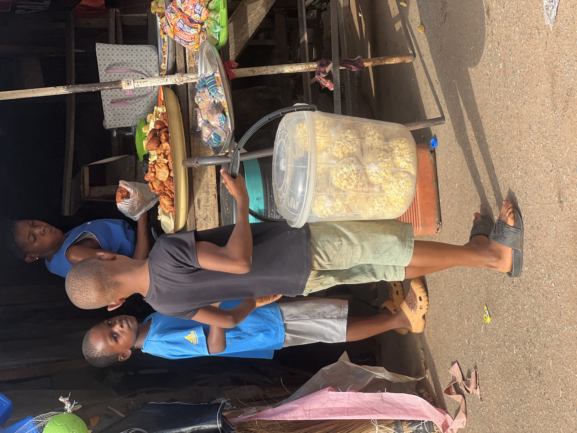The image, taken in Obosi, Nigeria, captures an outdoor street vending scene on a bright, sunny day. Three young males are present around a makeshift stall constructed from wooden poles and metal frames.
In the foreground, a boy wearing a blue t-shirt with the "Adidas" logo looks directly at the camera with a neutral expression. Next to him, another boy in a black t-shirt and khaki shorts stands, holding onto the frame of a vending cart. He is wearing one dark slipper and has one bare foot. Partially obscured behind the stall is a third boy wearing a blue sleeveless top.
The vending setup features a large clear plastic container filled with numerous small, individually packed bags of popcorn or a similar snack. Adjacent to this, a long metal tray holds a variety of fried foods, including plantain slices and what appear to be fried pastries or yam. Another section of the stall displays packaged snacks in colorful wrappers. A small clear plastic bag containing more fried items is also visible.
The ground is paved, and long shadows cast by the people and the stall indicate bright daylight, suggesting either morning or late afternoon. A pink fabric, a broom, and other assorted items are visible near the base of the stall. The scene portrays typical informal commerce in a roadside setting.