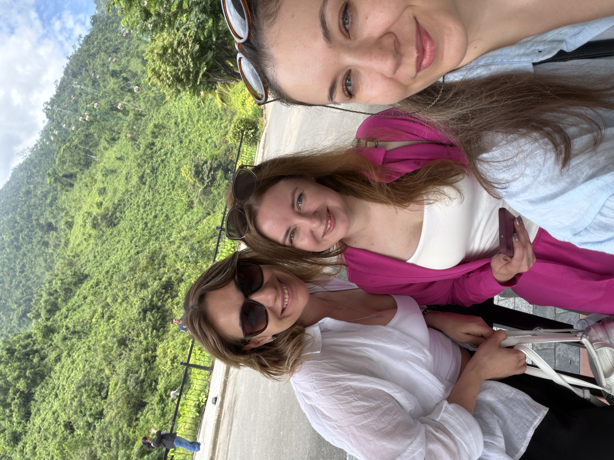 Three women are taking a selfie outdoors in front of a lush, green mountainside. The woman on the far left is wearing sunglasses, a white unbuttoned shirt over a white tank top, and black pants. She is smiling and holding a white purse. The woman in the middle has long auburn hair, is wearing a bright pink jacket over a white top, and is looking at her phone. The woman on the far right is wearing a light blue shirt and also smiling for the camera. In the background, the mountains are densely covered with vegetation, and cable cars are visible ascending the slopes, suggesting the location is Ba Na Hills in Đà Nẵng, Vietnam. The ground in the foreground appears to be paved.