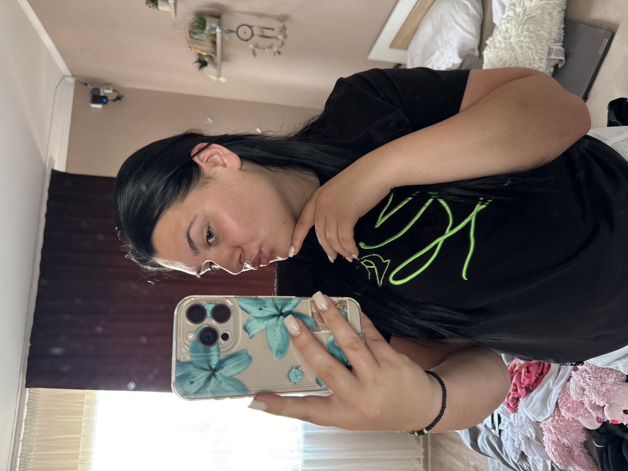 A woman with long, dark hair is depicted in a mirror selfie, standing in what appears to be a bedroom in Rogosh, Bulgaria. She is wearing a black t-shirt with a neon green linear design and a black braided bracelet with green accents on her left wrist. Her fingernails are painted a light opaque color. She holds a smartphone, encased in a clear cover adorned with teal floral patterns, in her right hand, while her left hand rests near her chin. Her lips are pursed in a 'kiss' expression. The background features a light pink/beige wall on the right, with two small floating shelves holding potted plants and decorative items, and a dreamcatcher hanging below them. High on the right wall, a small dark electronic device with a blue indicator light is mounted, with visible wiring. To the left, a window with light-colored sheer curtains is visible behind darker, possibly brown, drapes. A white ceiling with molding runs across the top. In the lower right, part of a bed with a white headboard and white bedding is visible, alongside a white fluffy pillow and a silver/grey laptop (Lenovo). The lower-left corner shows a pile of clothes and several pink stuffed animals.