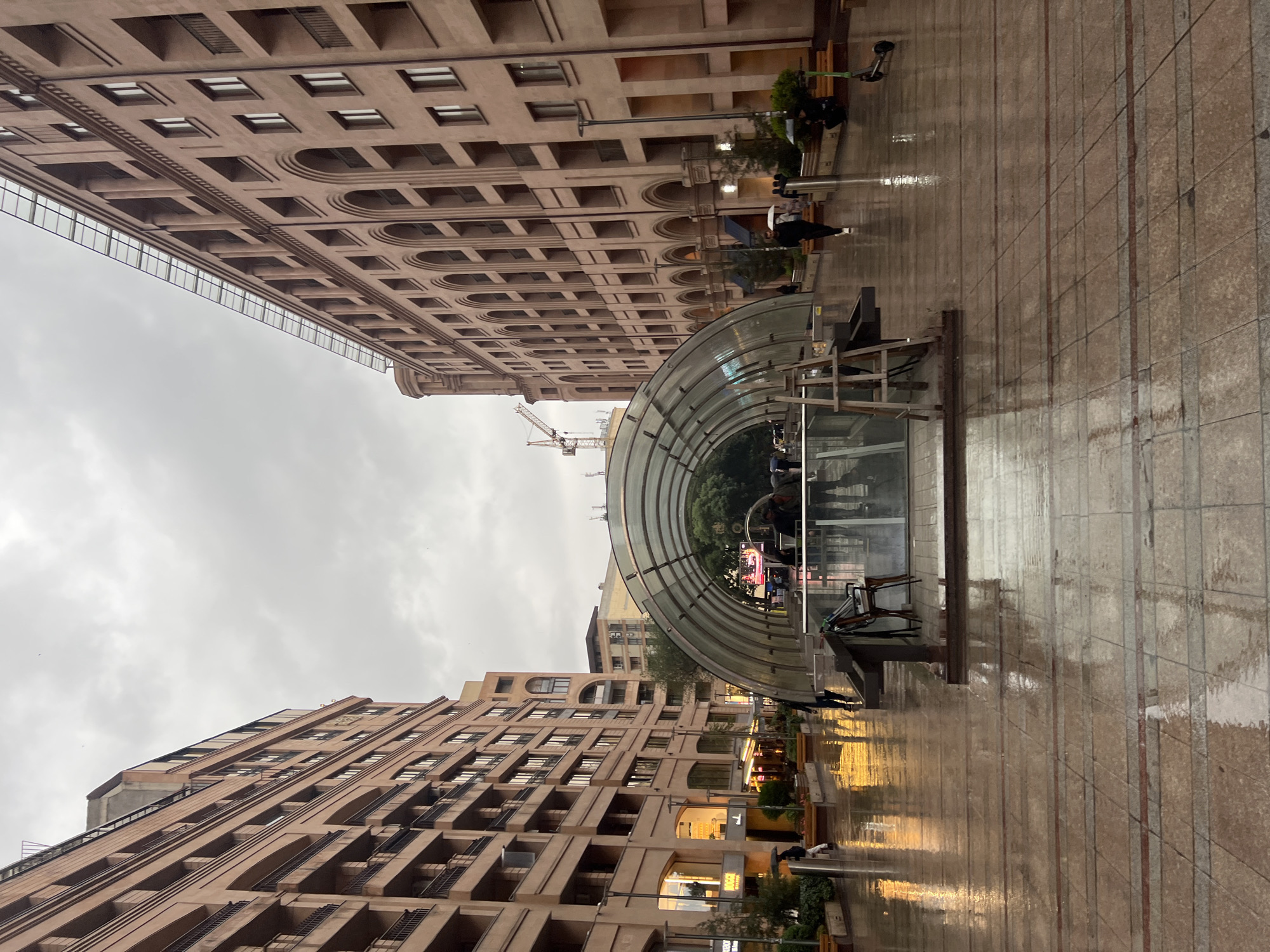 The image depicts a street scene in Yerevan, Armenia, likely Republic Square based on the architectural style of the buildings. Two large buildings flank the street, each featuring multiple stories and arched windows. The ground is paved with wet, reflective tiles. A semi-circular, transparent structure sits in the foreground, potentially a metro entrance or some form of public shelter. The sky is overcast. A construction crane is visible in the distance. People are sparsely distributed along the street.