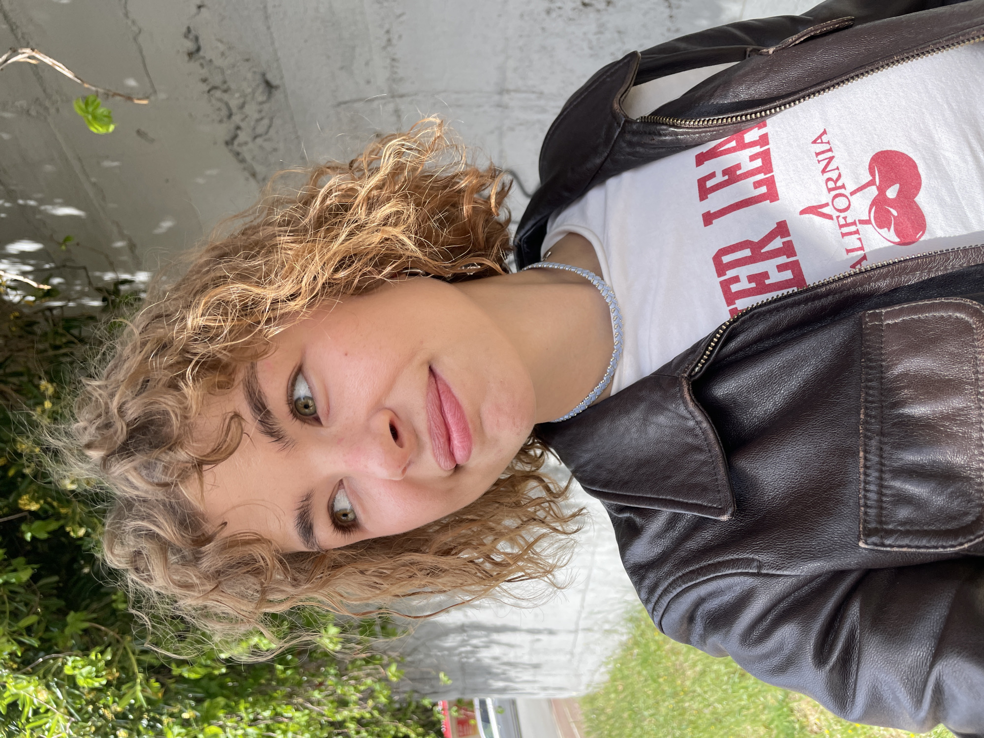 A young woman with curly hair is wearing a brown leather jacket over a white T-shirt. The shirt has the words "Cherry LA California" printed in red. She also wears a blue and white beaded necklace. The background consists of a concrete wall, greenery, and a street.