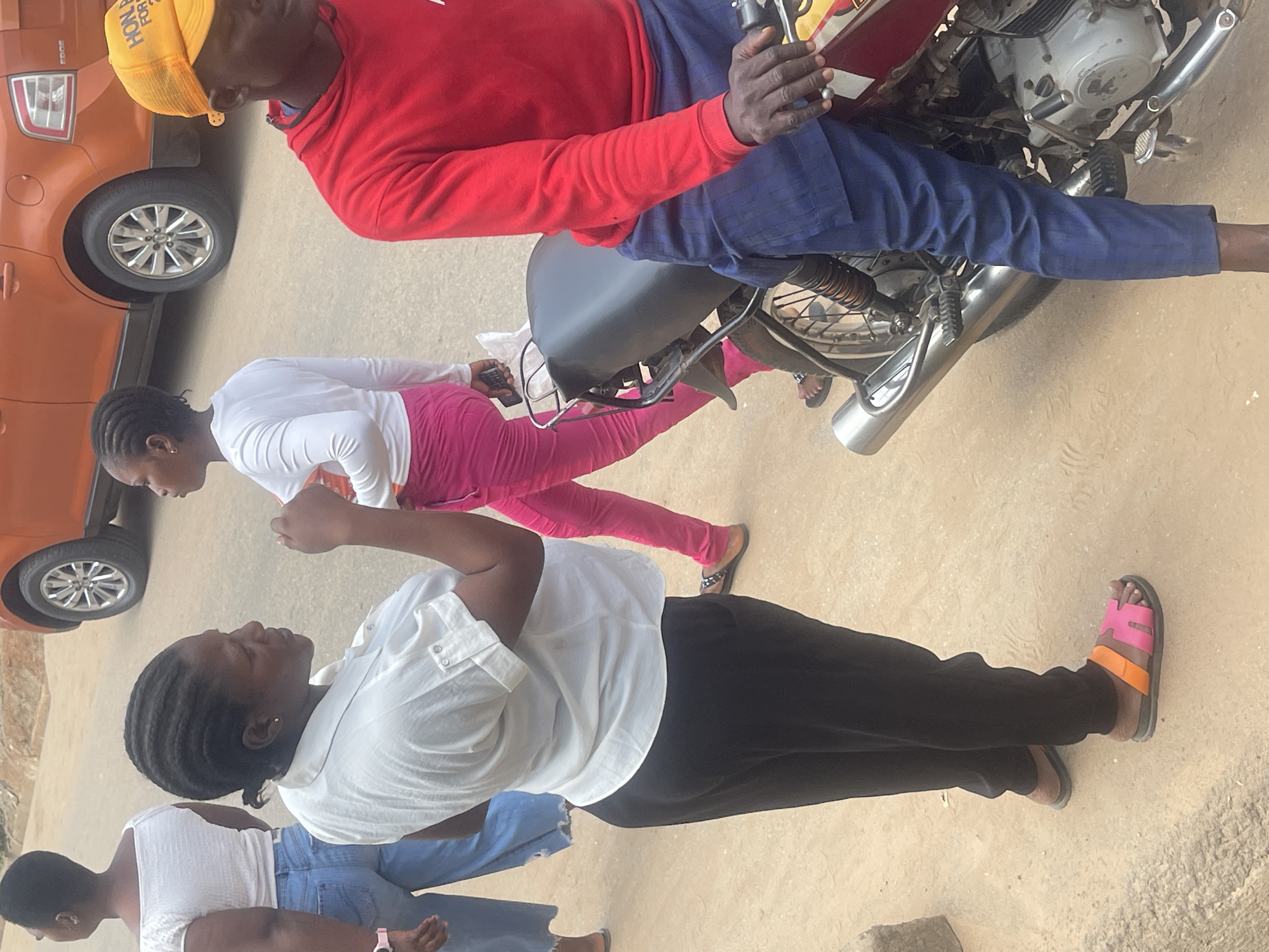 The image shows a group of people in what appears to be a street scene, possibly in Abuja, Nigeria. In the foreground, a woman in a white shirt and black pants is the primary subject, facing away from the camera with her head turned to the side. She is wearing orange and pink sandals. Behind her, another woman in a white top and blue jeans is also visible. Further back, a person in a red long-sleeved shirt and blue pants is seated on a motorcycle. This person is wearing a yellow cap with the partial text "HON FOR" visible. Another individual in a white long-sleeved shirt and bright pink pants is standing near the motorcycle. The background shows an orange car. The lighting suggests it is daytime, and the ground appears to be dirt or gravel. The overall impression is a casual, everyday scene with people gathered around a motorcycle.